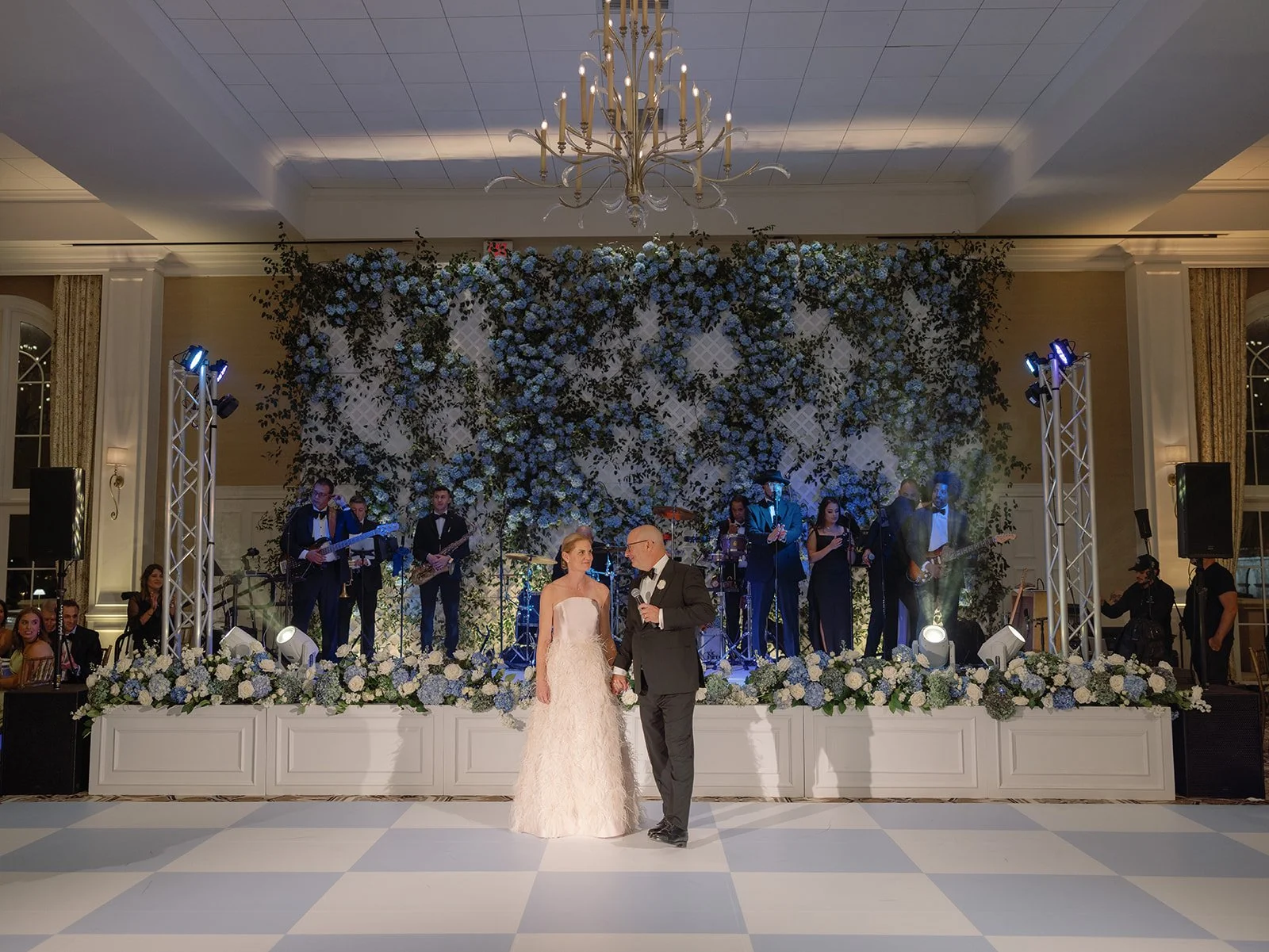 A bride and groom holding hands during their wedding reception dance in front of a live band with a floral backdrop. The room features a chandelier and decorated tables, with guests seated and musicians playing instruments.