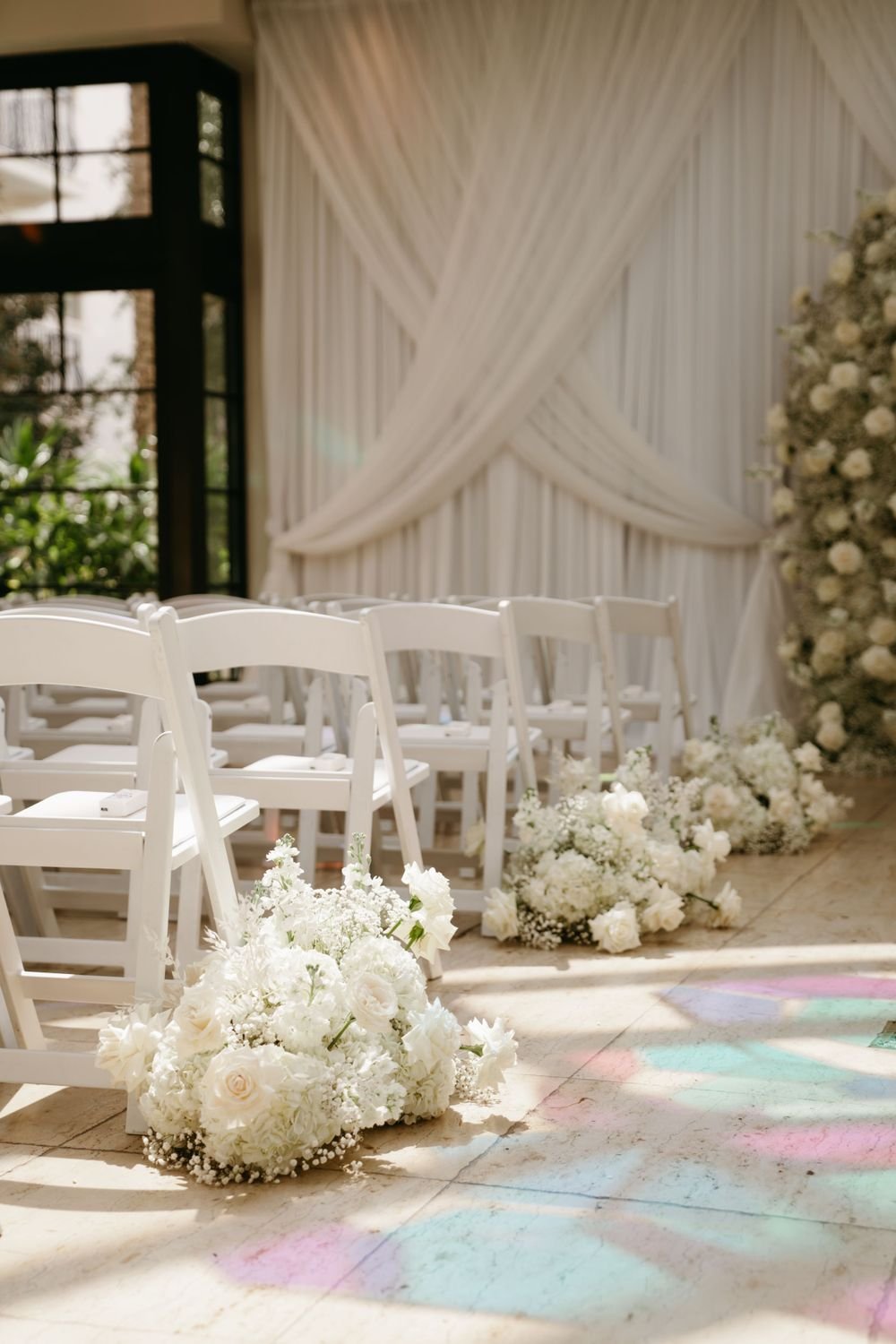 Wedding ceremony setup with white chairs decorated with white floral arrangements along the aisle, white drapery backdrop with a large floral arrangement nearby, and a window allowing natural light to illuminate the scene.