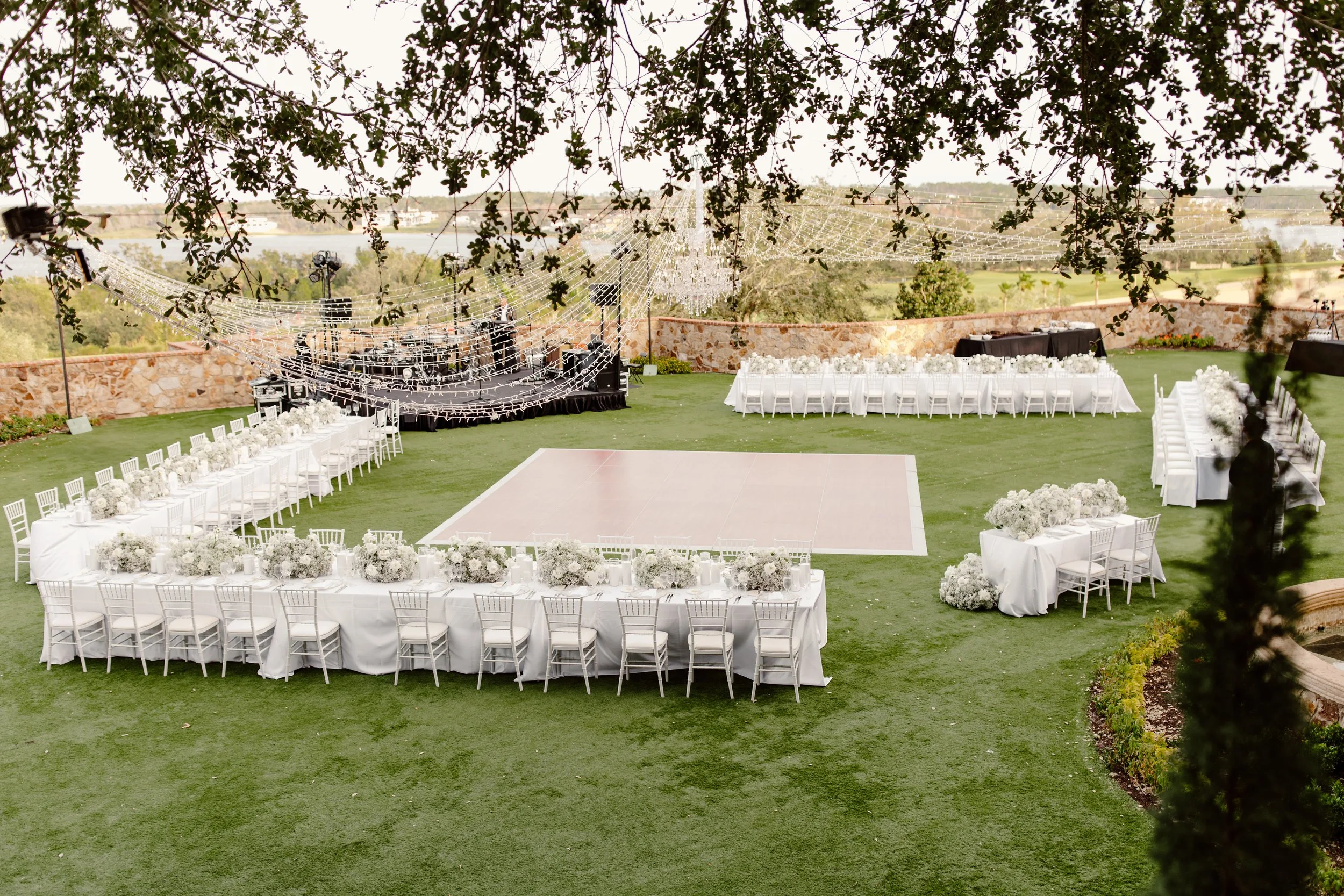 Outdoor wedding setup with a dance floor, long reception tables with white tablecloths and floral centerpieces, surrounded by white chairs. Overhead, there are string lights and decorative chandeliers hanging, with a stone wall and scenic landscape i
