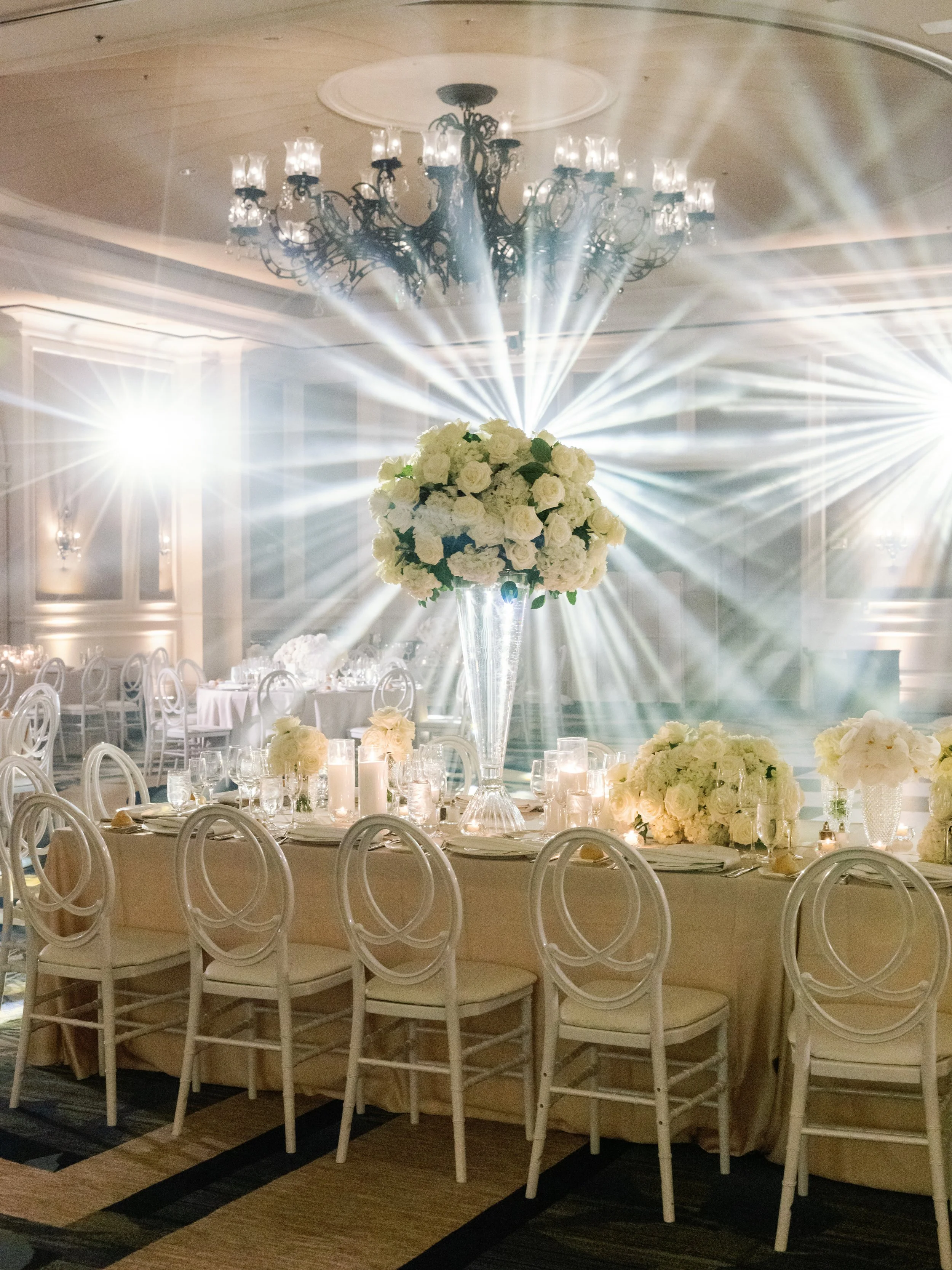 Elegant banquet hall featuring a tall floral centerpiece with white roses and hydrangeas, surrounded by candles and glassware on a round table. In the background, a chandelier with multiple lights hangs from the ceiling, and bright light rays emanate