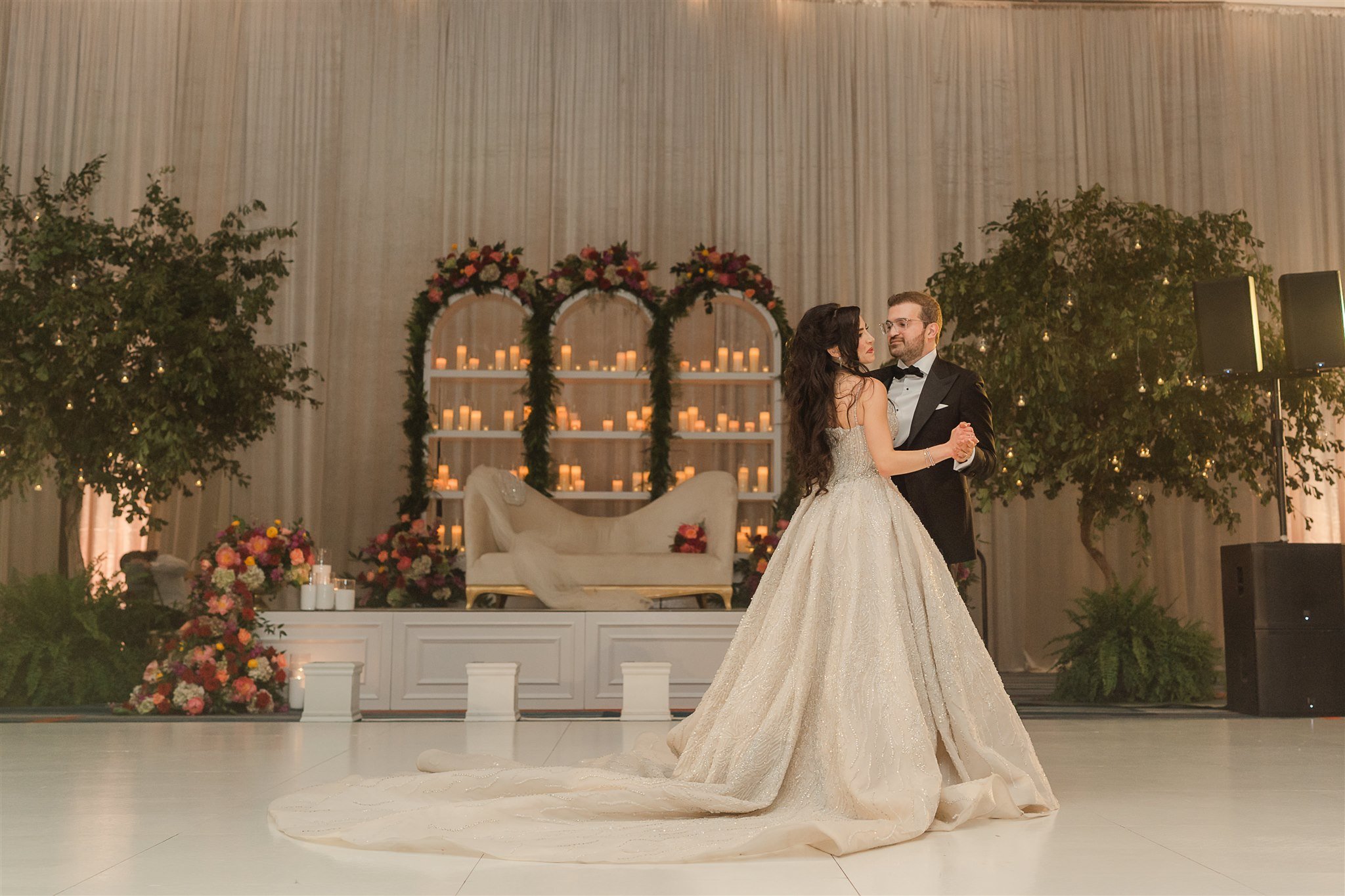 A bride and groom are dancing during their wedding reception inside a decorated venue with floral arrangements, candles, and greenery.