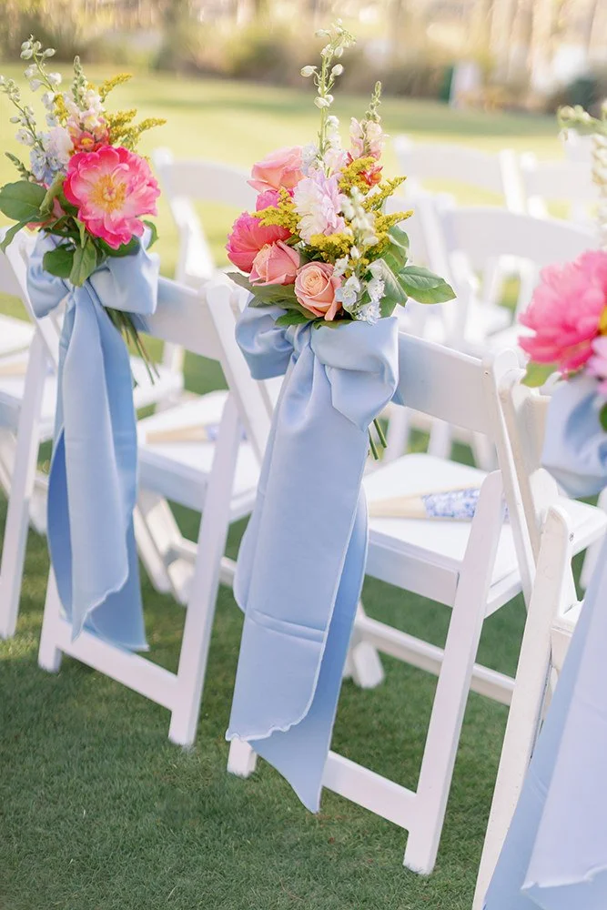 White folding chairs decorated with pink and white floral arrangements and light blue ribbons, set up outdoors for an event.