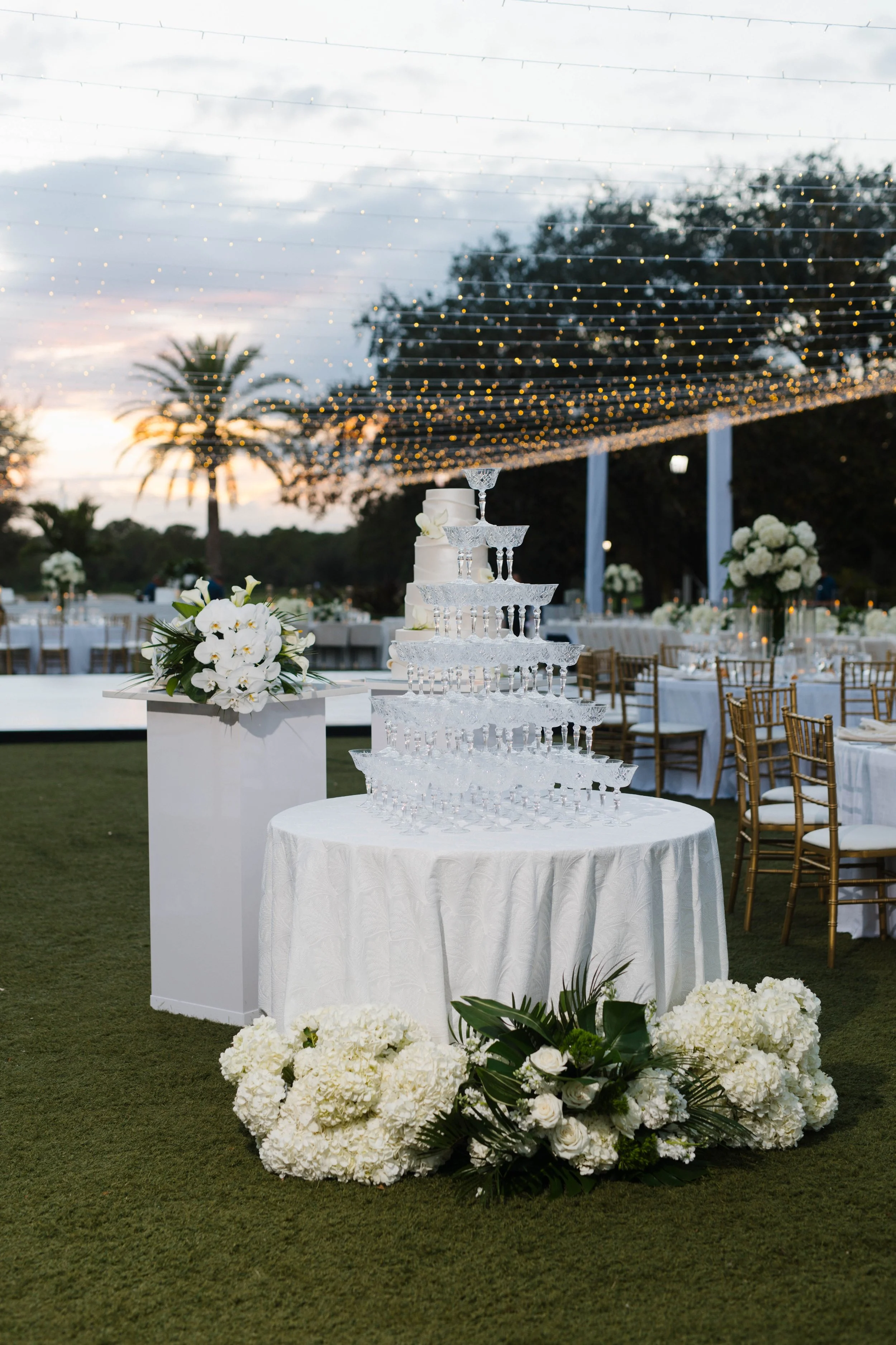 Elegant outdoor wedding reception setup with a tiered champagne tower, floral arrangements with white flowers, and twinkling string lights overhead at sunset.