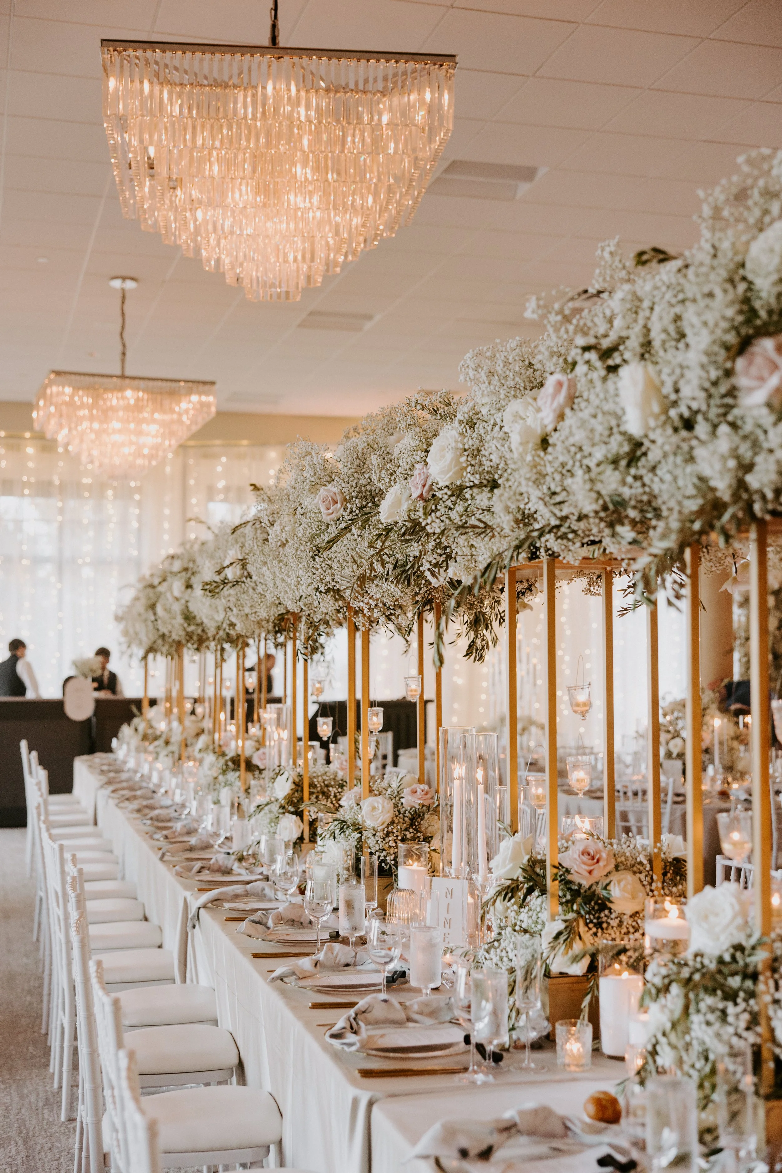 Elegant wedding reception table decorated with white flowers, candles, and tall floral centerpieces, with chandeliers hanging from the ceiling.