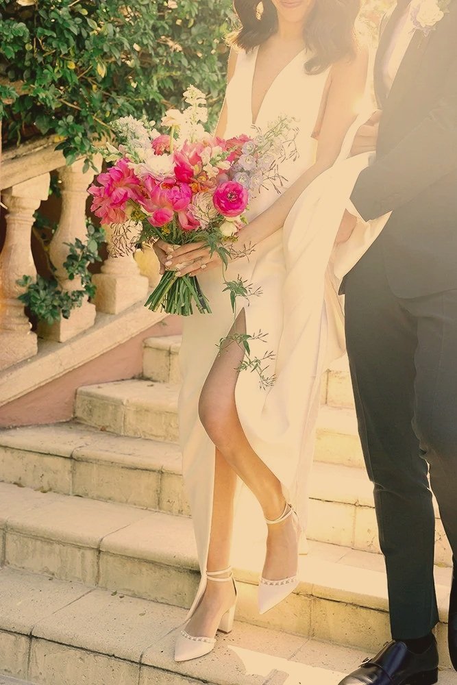 Close-up of a bride holding a vibrant bouquet, wearing a white dress with a high slit, standing on stone steps with a groom partially visible beside her.
