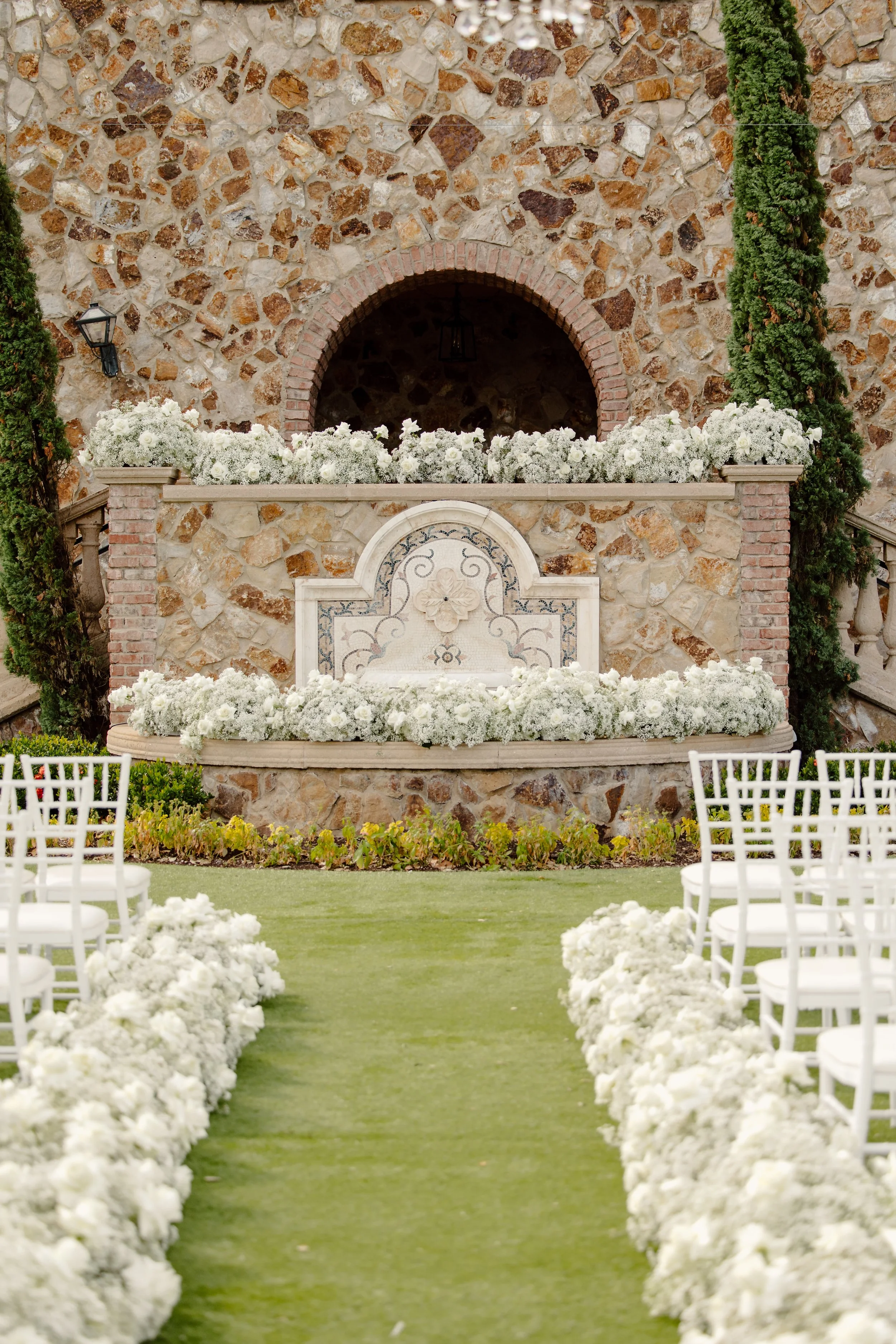 Outdoor wedding aisle with white floral arrangements on either side, leading to a stone wall with an arched niche decorated with more white flowers, flanked by two tall green trees.