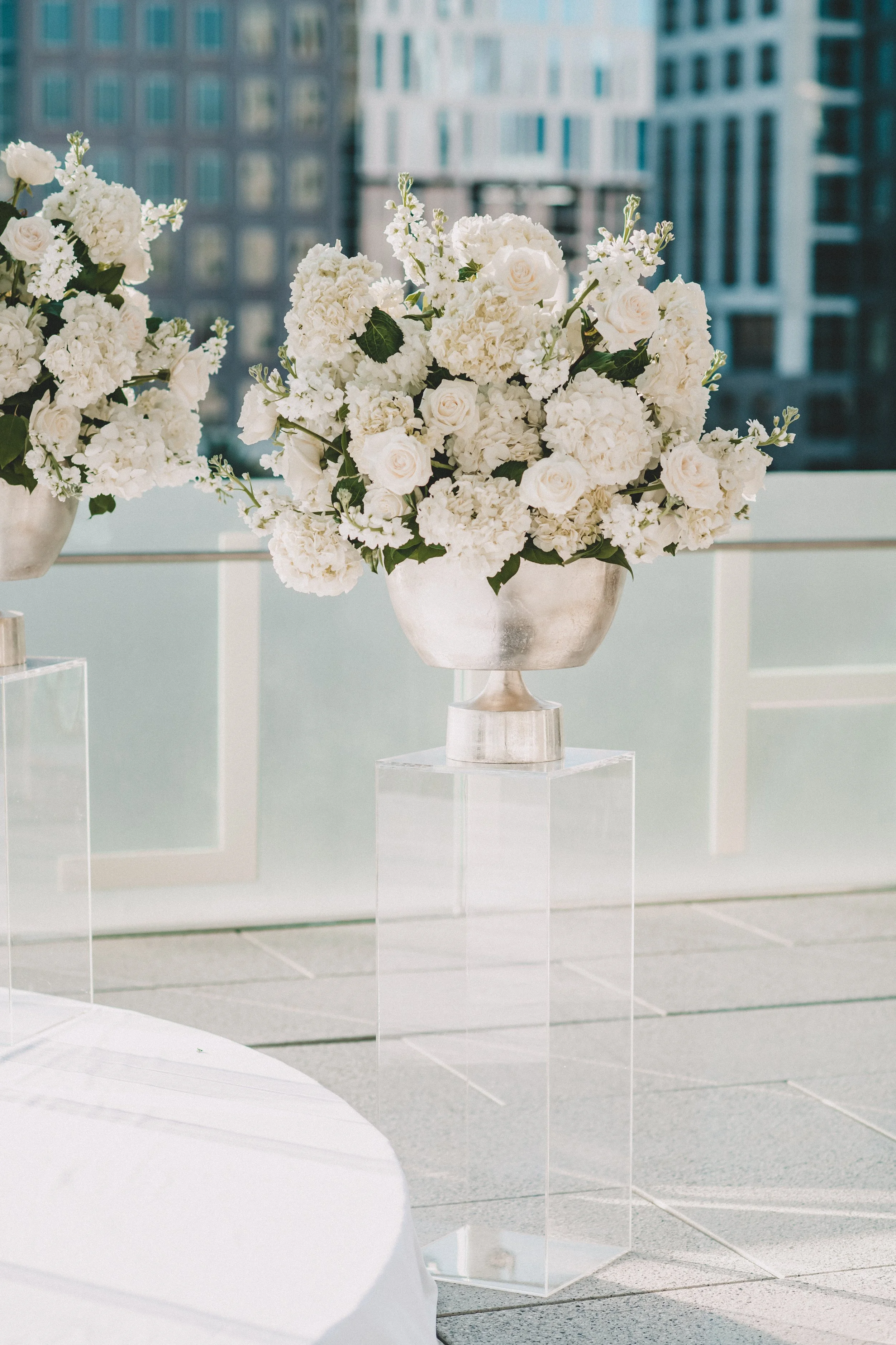 A metallic vase with white flowers, including roses and hydrangeas, is displayed on a white stand against a cityscape with tall buildings through a window.