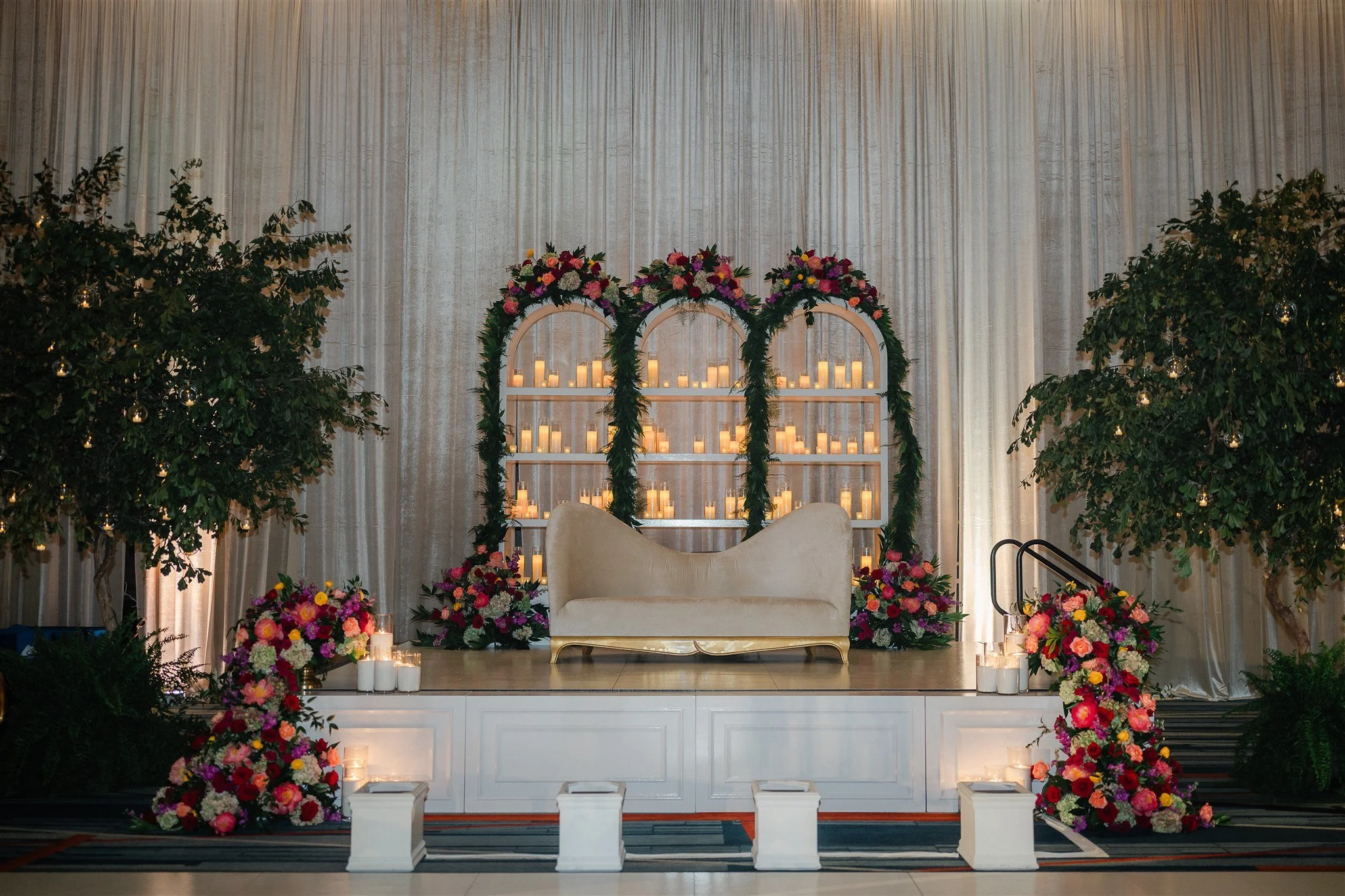 A wedding altar decorated with large colorful flower arrangements and a white sofa in front of a background with candles and draped curtains.