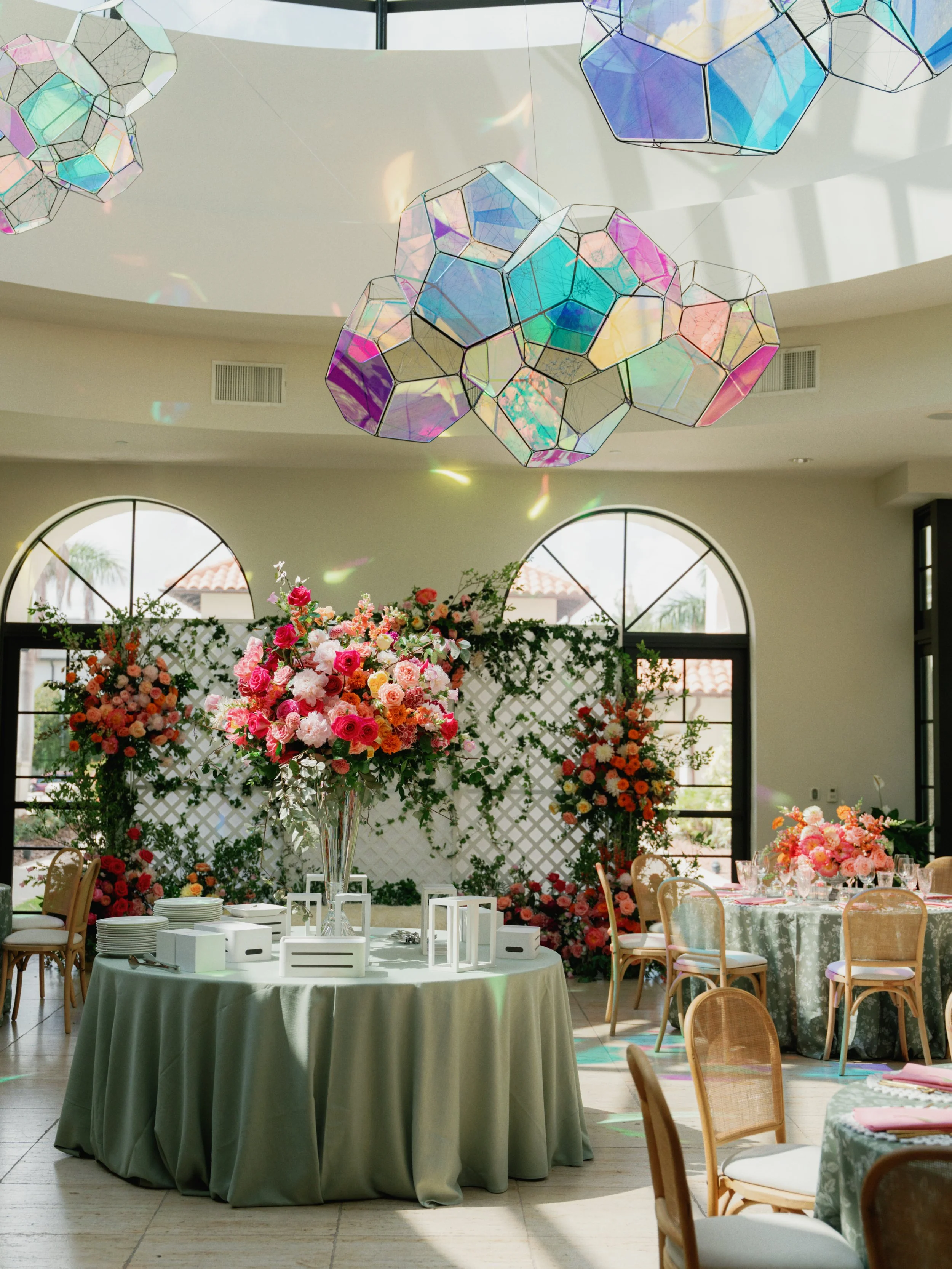 Elegant indoor party setup with round tables covered with floral tablecloths and decorated with large flower arrangements. The background features large floral decorations and geometric pendant light fixtures hanging from the ceiling. The room is wel