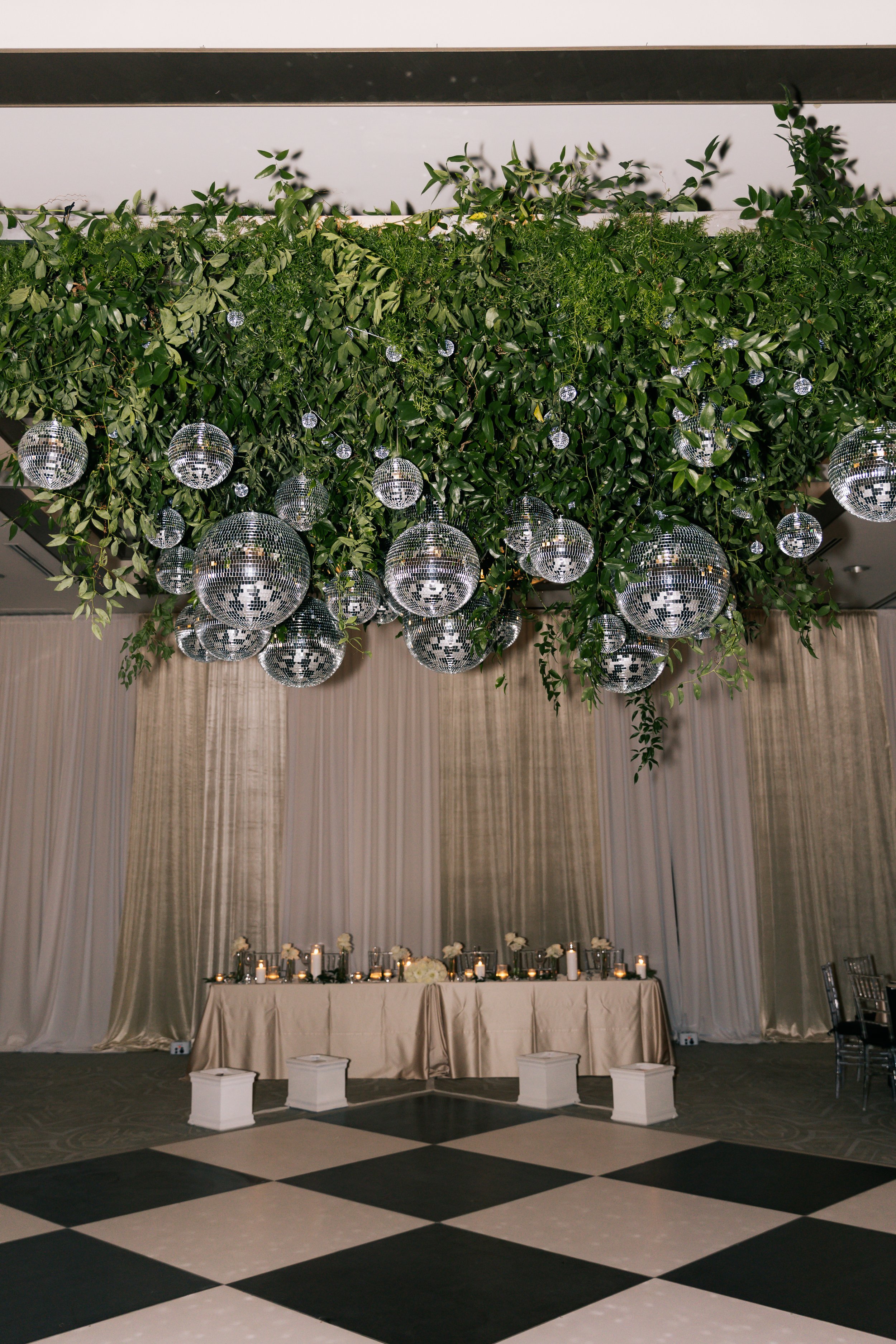 Event space decorated with a large green leafy foliage installation on the ceiling, adorned with numerous disco ball ornaments. Below, a long table is decorated with candles, flowers, and white drapery, set against a backdrop of white curtains and dr