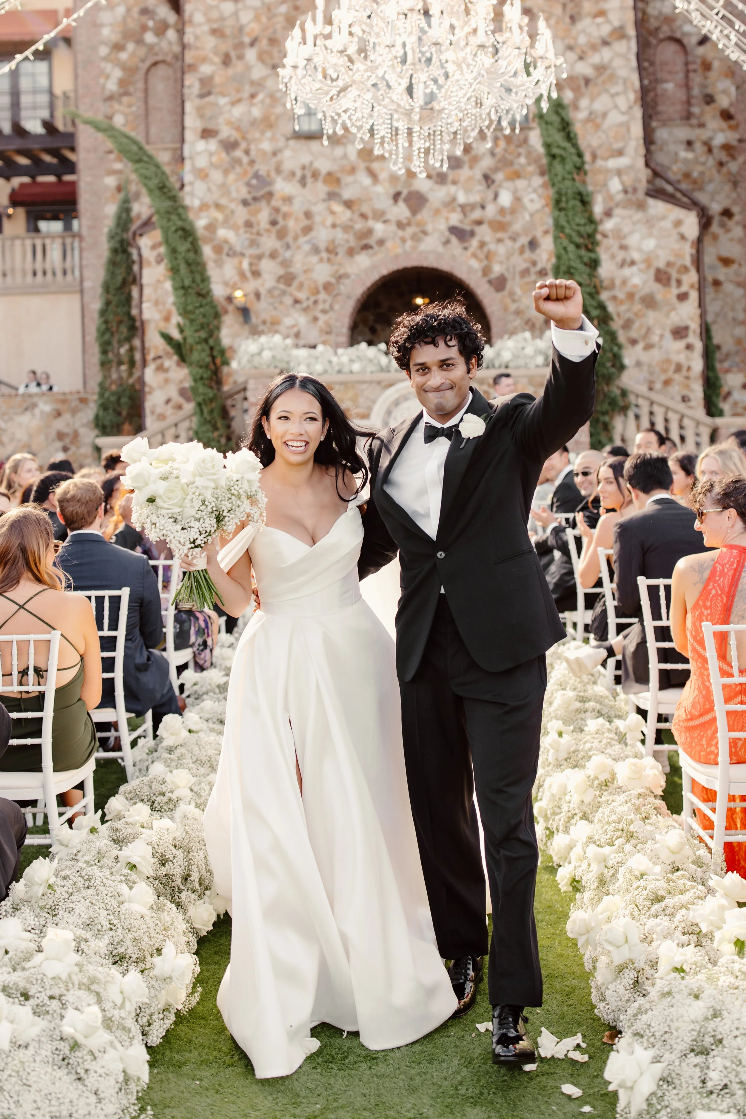 A bride and groom walking down the aisle after their wedding ceremony, surrounded by guests and floral decorations, with a large chandelier hanging above, in an outdoor venue with a stone building in the background.