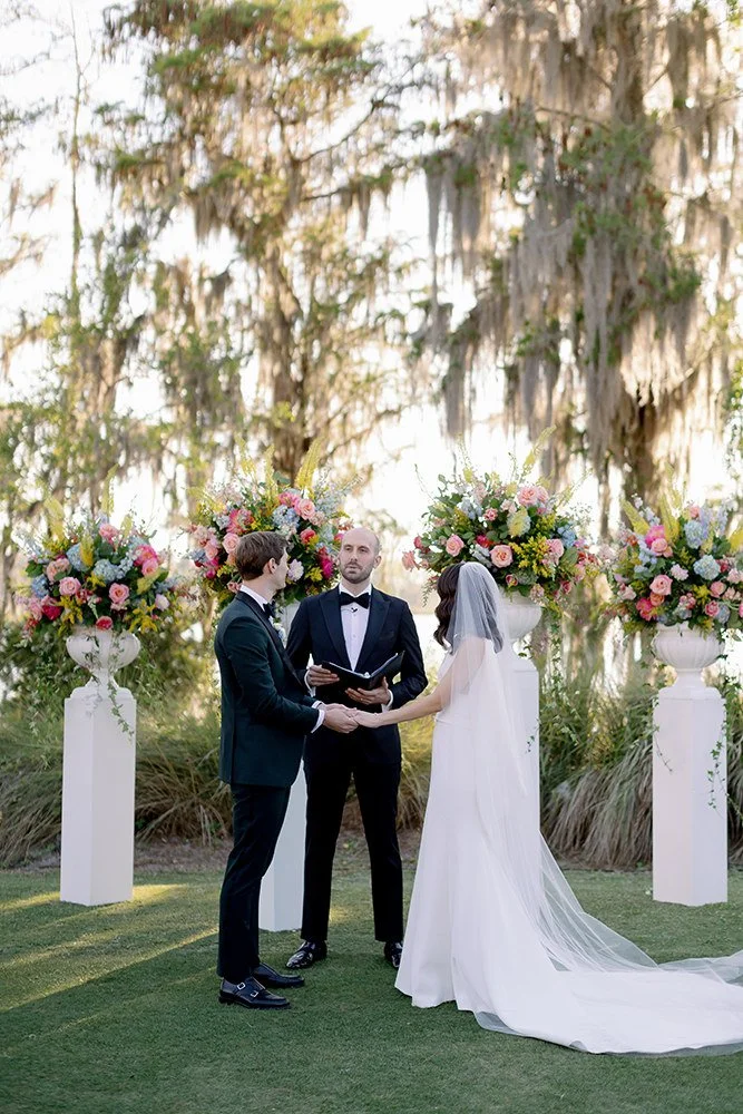 A wedding ceremony outdoors with a bride and groom holding hands and facing each other, standing in front of an officiant surrounded by large floral arrangements on white pedestals, with trees in the background.
