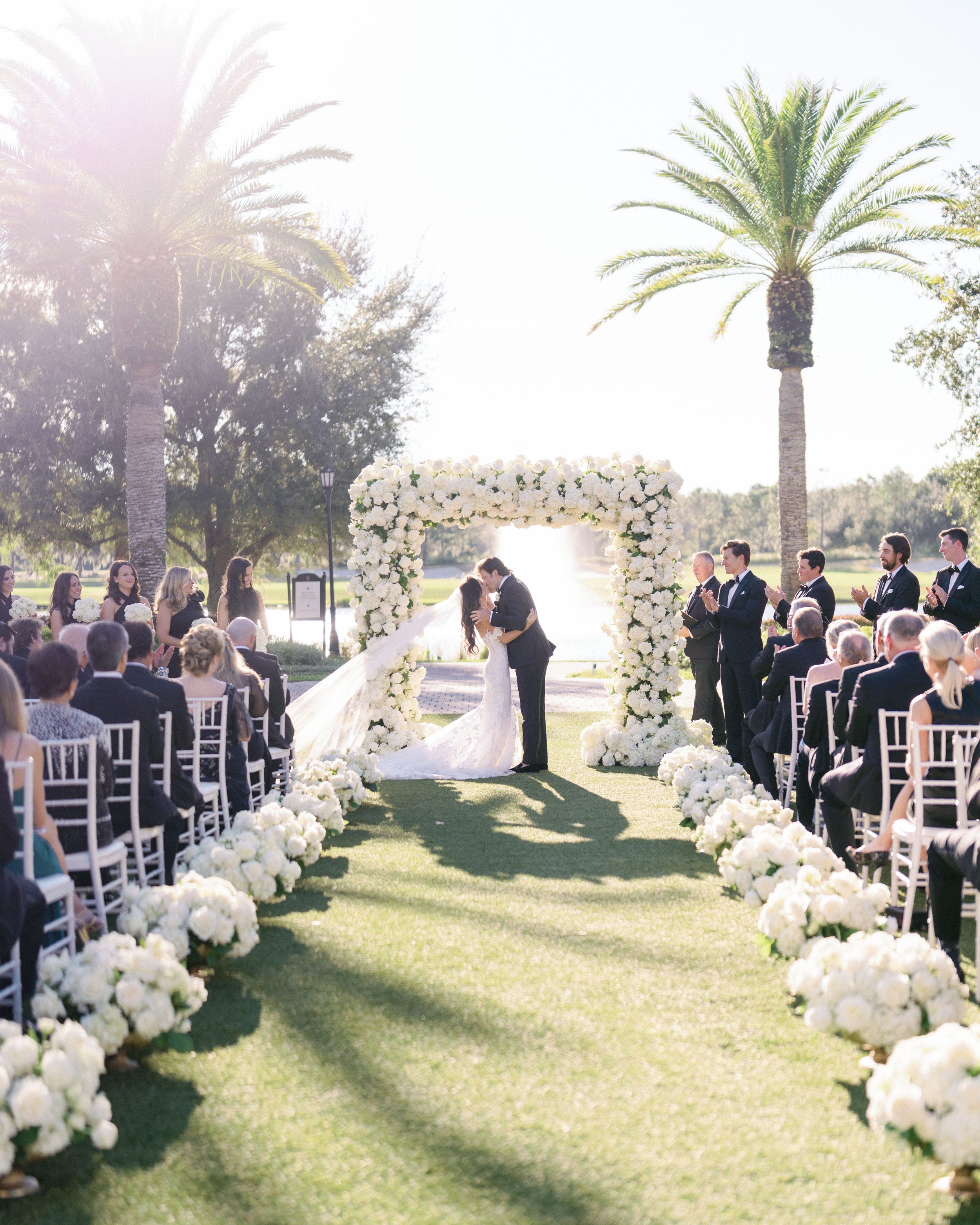 A wedding ceremony outdoors with a couple standing under a floral arch, surrounded by guests seated on white chairs, with palm trees and a water body in the background.