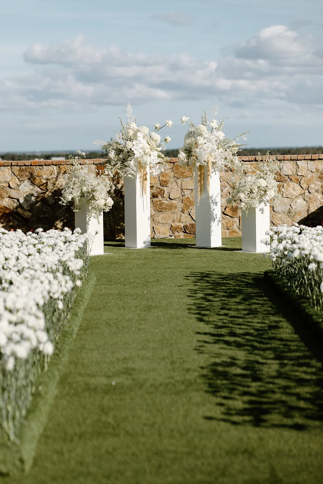 Wedding aisle with white flowers on either side and white floral arrangements on tall white stands at the altar, set outdoors against a blue sky with clouds and a stone wall in the background.