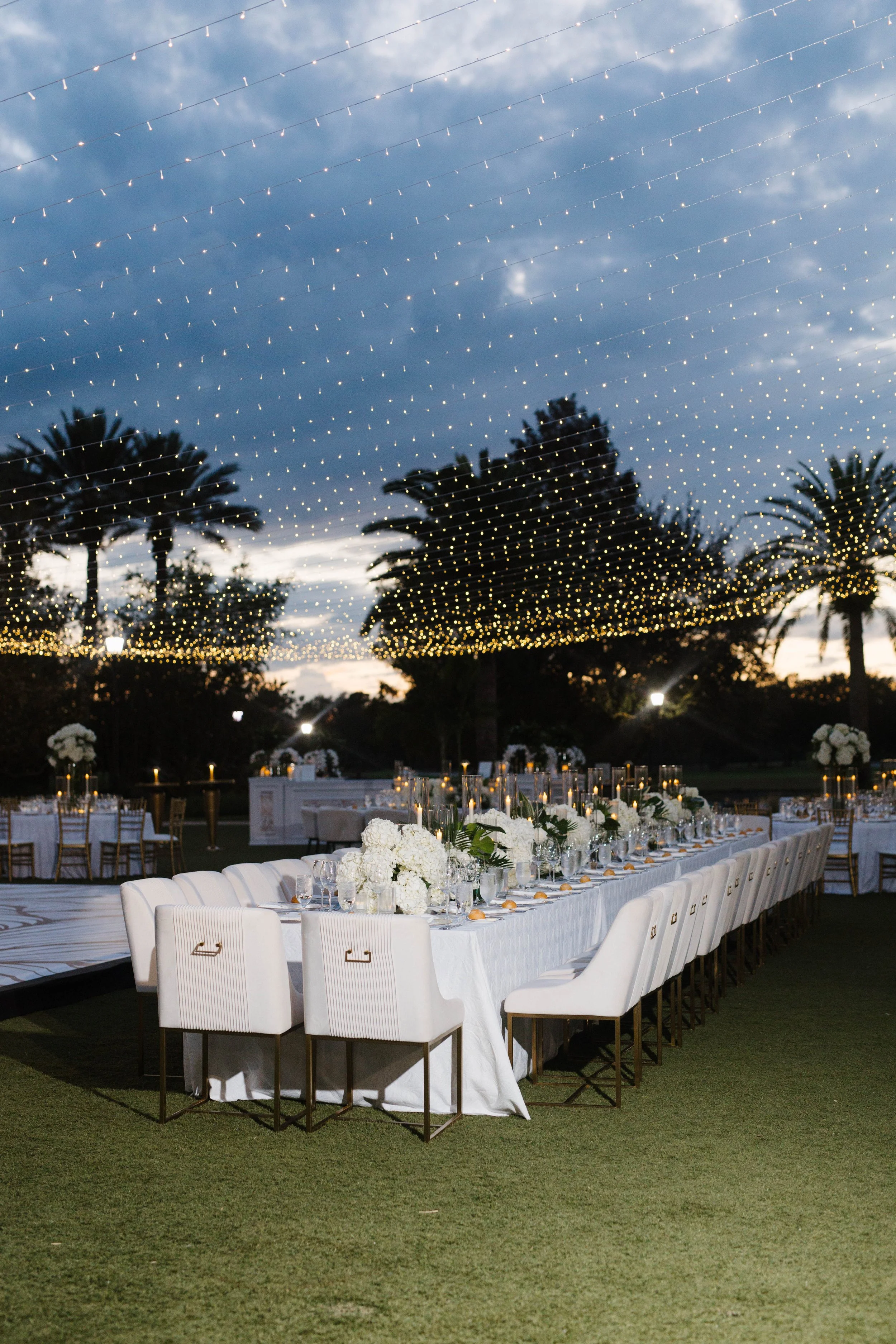 Outdoor wedding reception table with white tablecloth, floral centerpieces, and candles under string lights at dusk, with palm trees and cloudy sky in background.