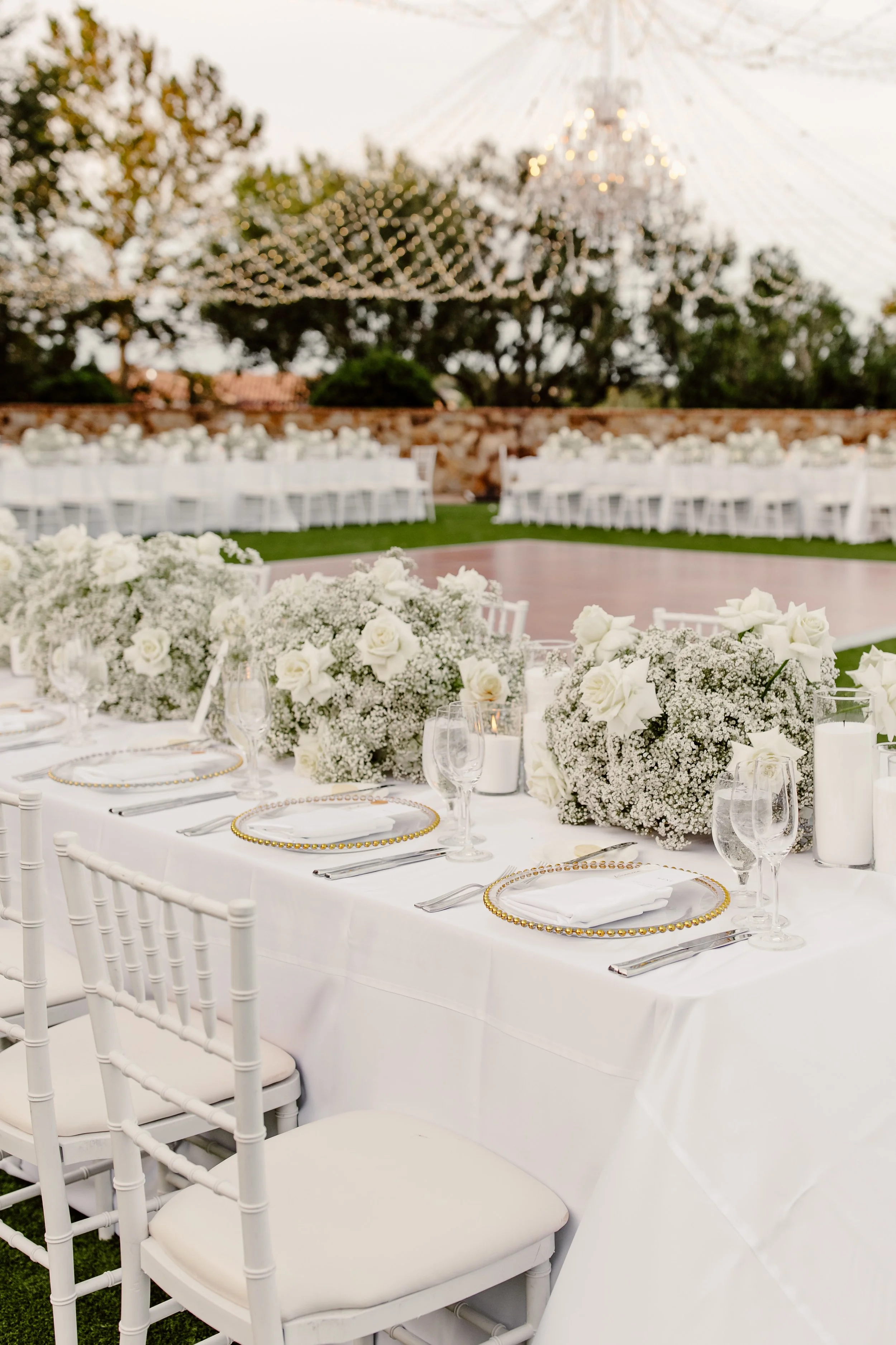 Wedding reception outdoors with white tables, floral centerpieces, and elegant table settings, with string lights and a chandelier in the background.
