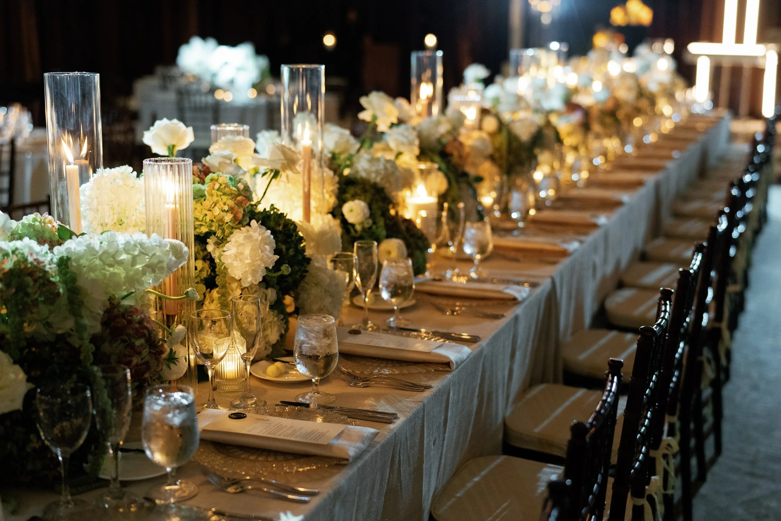 A long banquet table decorated with white floral centerpieces and candles, set for a formal event or wedding reception in a dimly lit room.