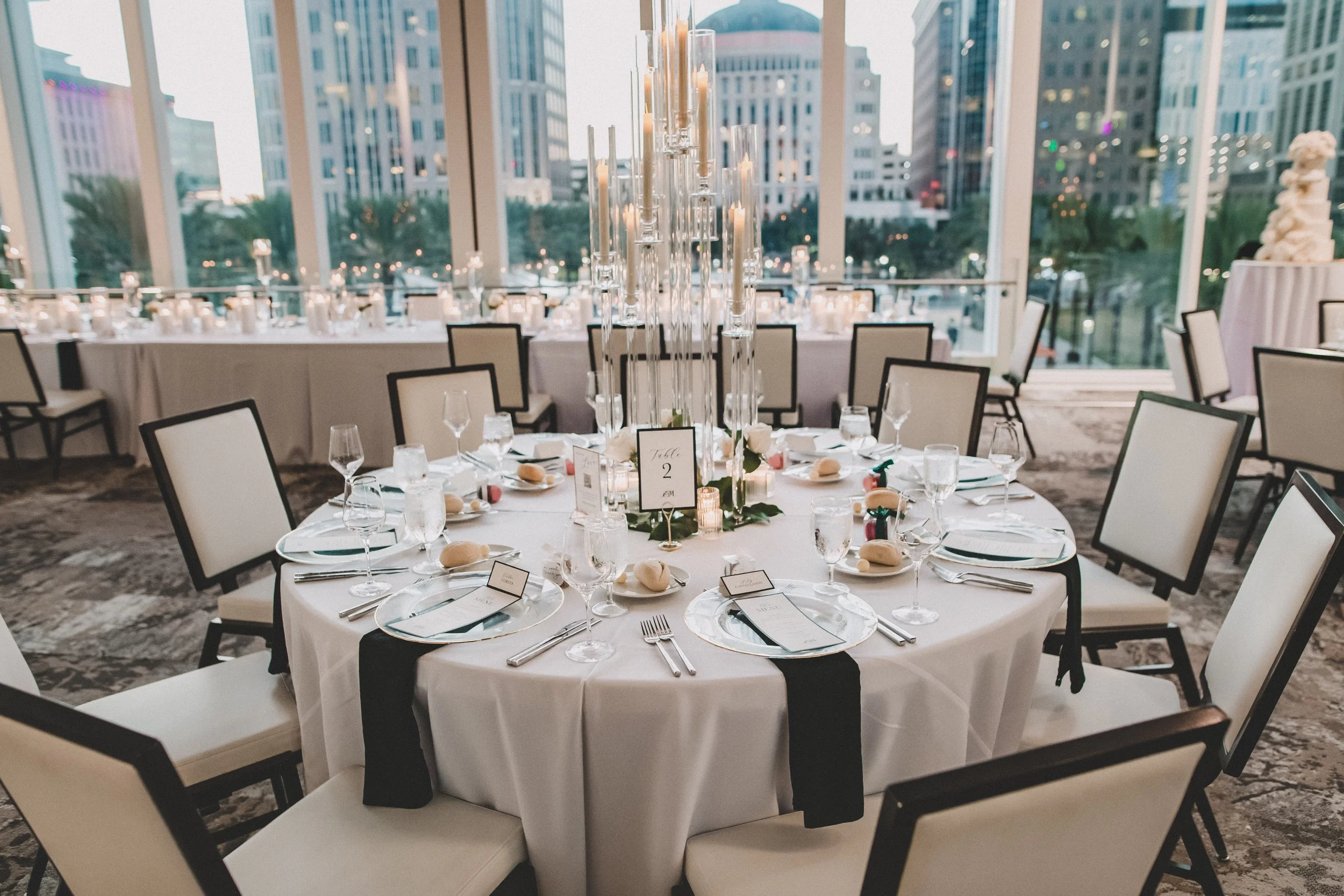 Round banquet table set for a formal event with white tablecloths, glassware, silverware, and bread rolls. Elegant centerpiece with tall glass candle holders and candles, surrounded by chairs in a modern venue with large windows showing a cityscape o