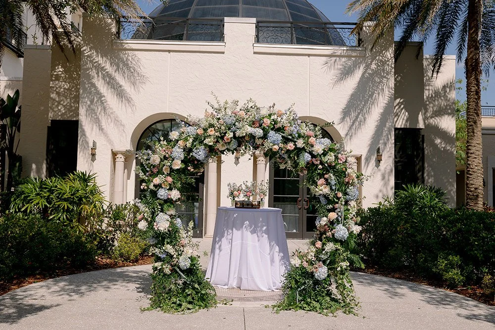 A floral arch decorated with pink, white, and blue hydrangeas and roses, surrounding a round table with a floral centerpiece, in front of an elegant white building with palm trees and glass dome roof.