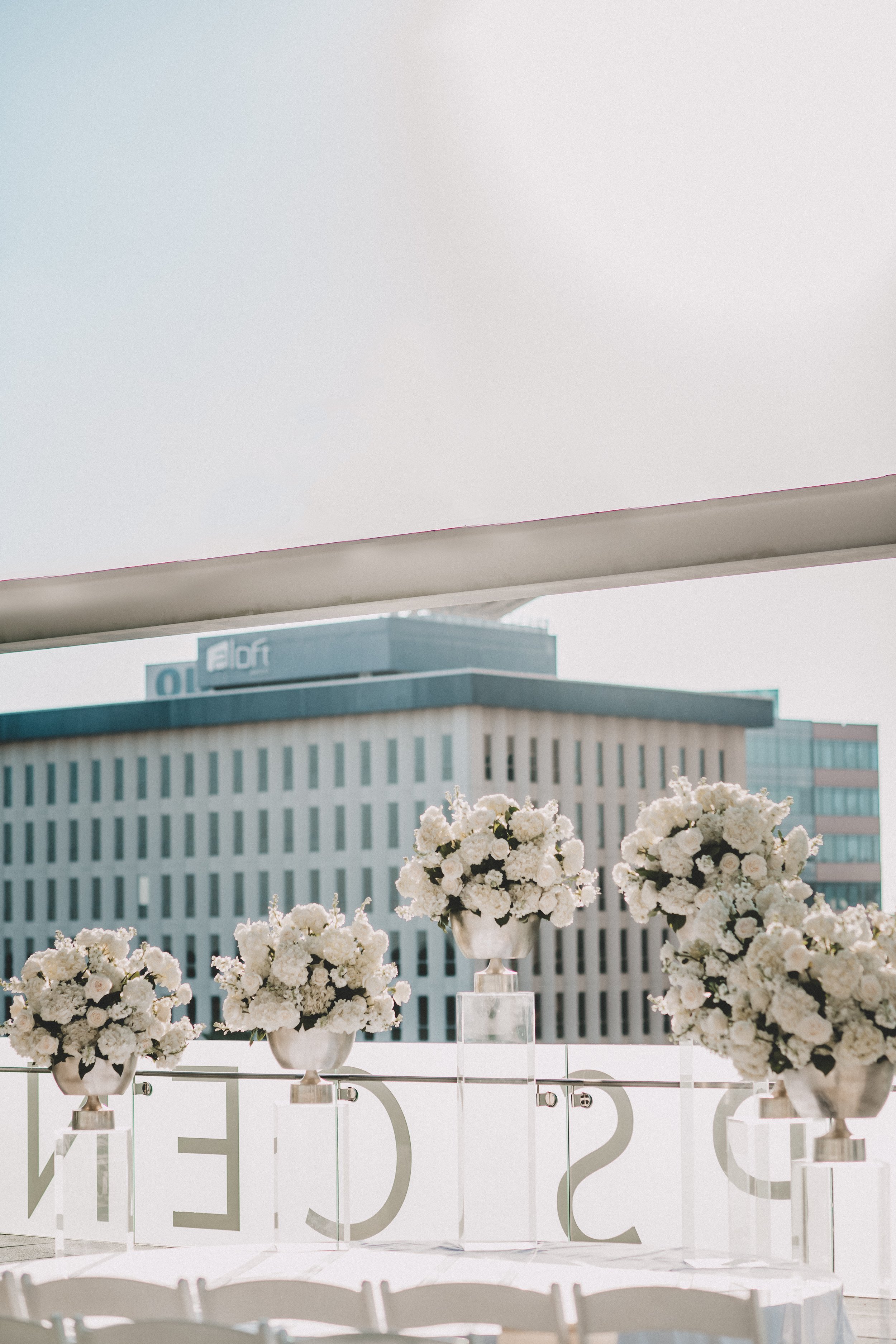 Elegant wedding or event setup on a balcony with white flowers in vases atop transparent pedestals, a white tablecloth, and large decorative letters spelling out a word behind the flowers, overlooking a cityscape with a modern office building.