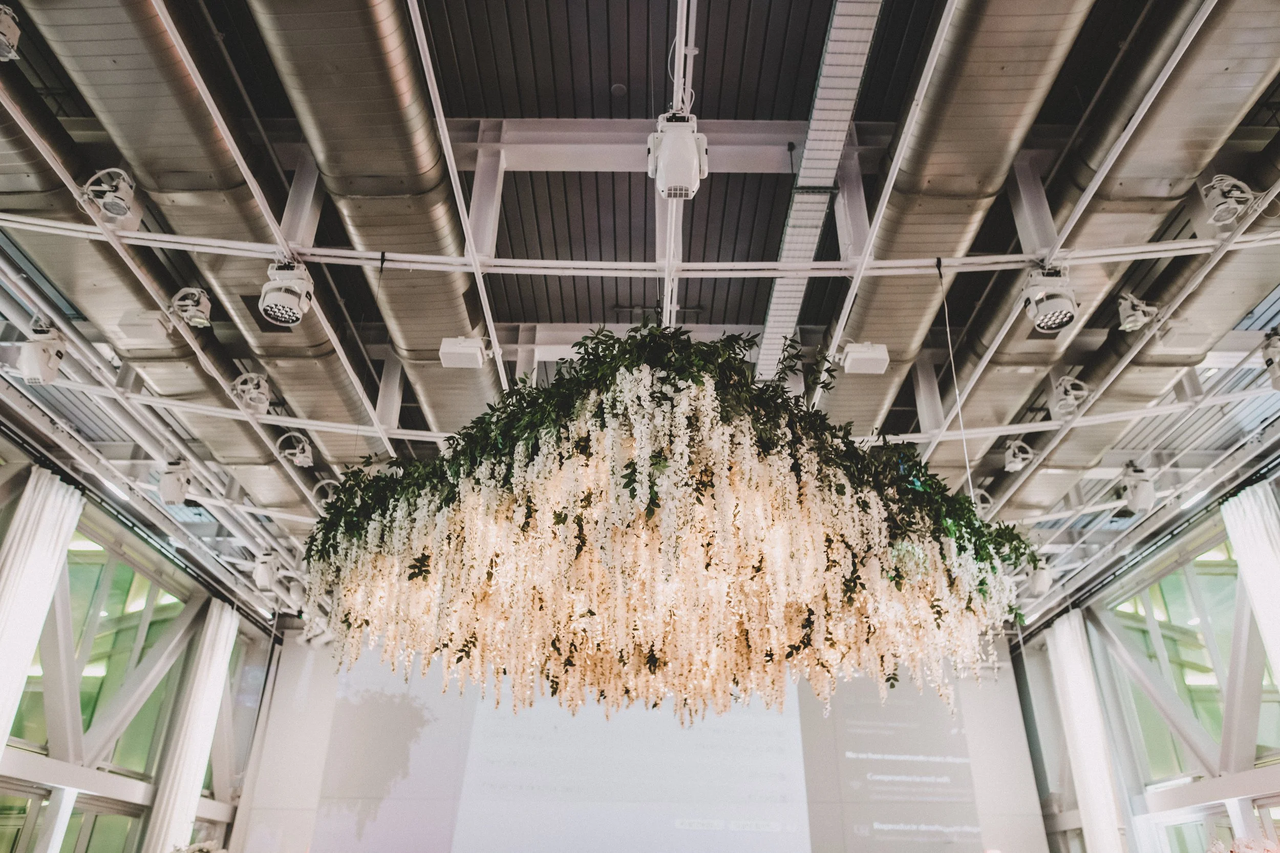 Hanging floral arrangement with white flowers and greenery in an indoor venue with a high ceiling, metal beams, and lighting fixtures.