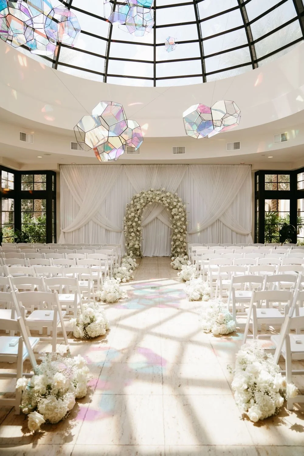 Elegant indoor wedding ceremony setup with white chairs, floral arch, and hanging geometric light fixtures in a bright room with large windowed walls.