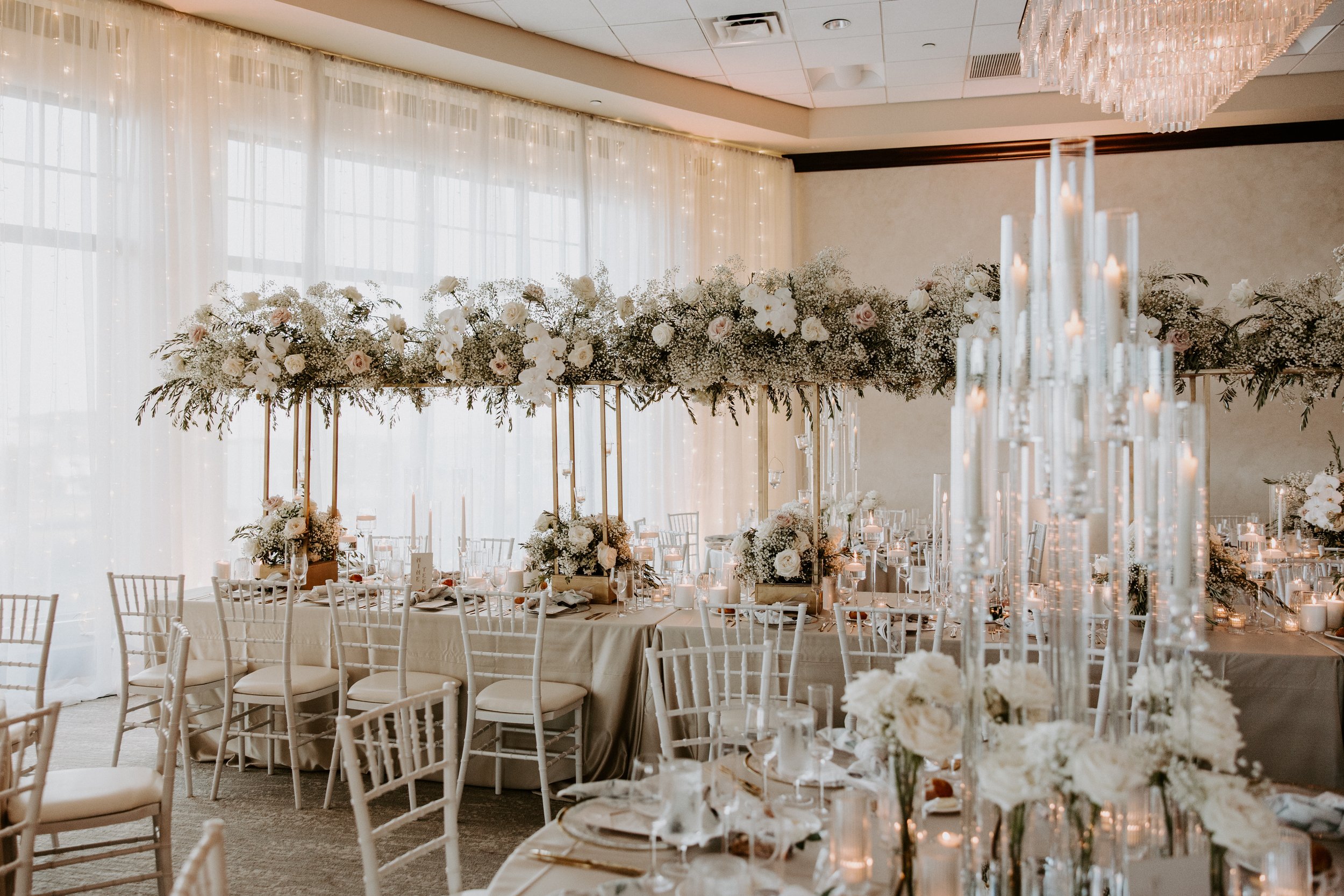 Elegant indoor wedding reception setup with long floral arrangements of white roses and baby's breath on elevated stands, surrounded by round and square tables with white chairs, tall candle holders with lit candles, and a backdrop of white sheer cur