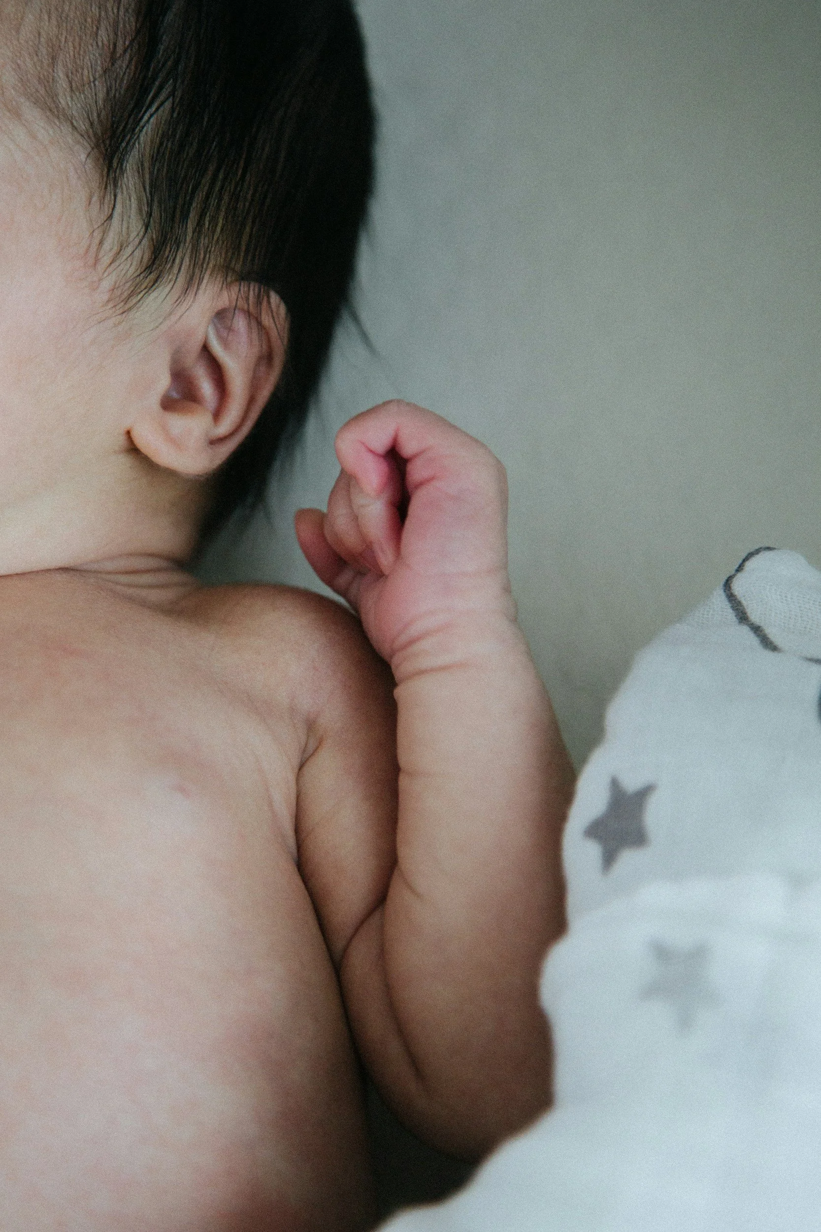 Close-up of a baby with a clenched fist, lying on a white blanket with star patterns, showing part of the head and shoulder.