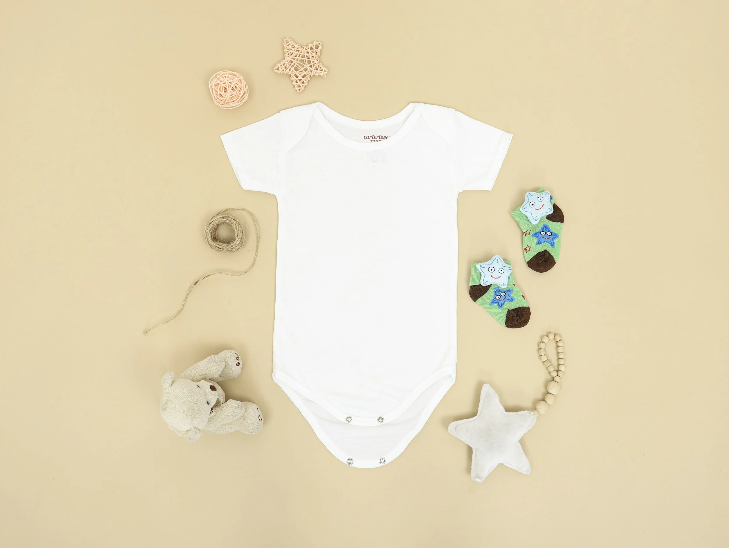 A white baby onesie, surrounded by a teddy bear, knitted toys, baby socks with cartoon faces, and various small decorative items on a beige background.