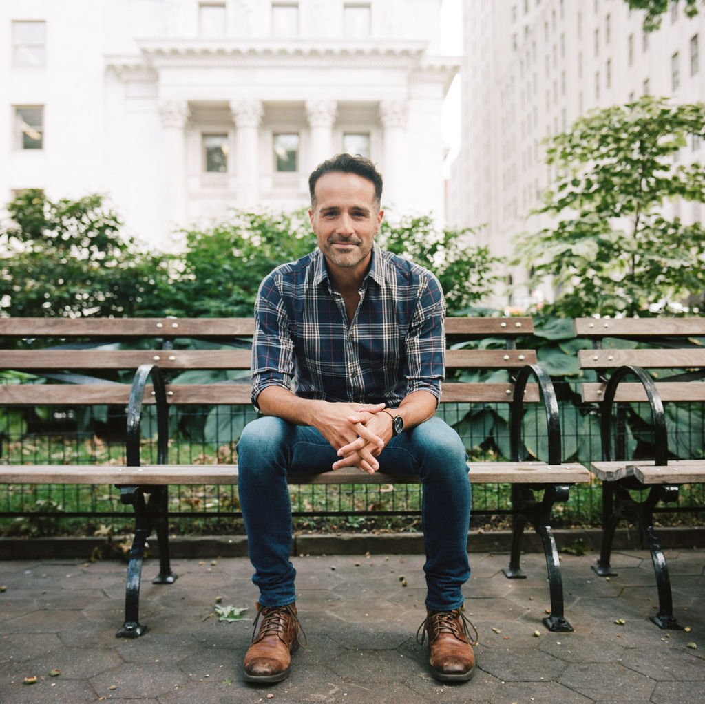 A man sitting on a park bench in an urban setting, with tall buildings and greenery in the background.