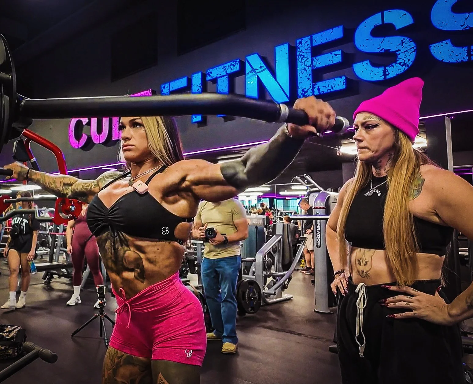 Two women with tattoos working out in a gym, one lifting a barbell and the other observing, with gym equipment and other people in the background.