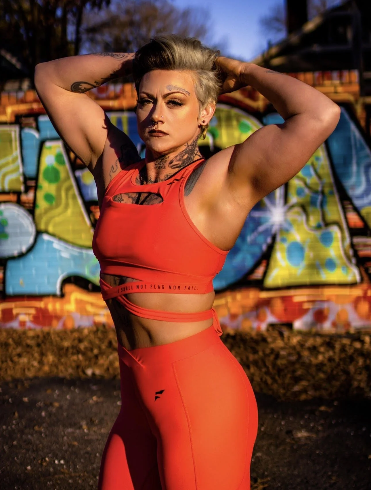 Woman with tattoos in red athletic wear posing outdoors in front of a colorful graffiti wall.