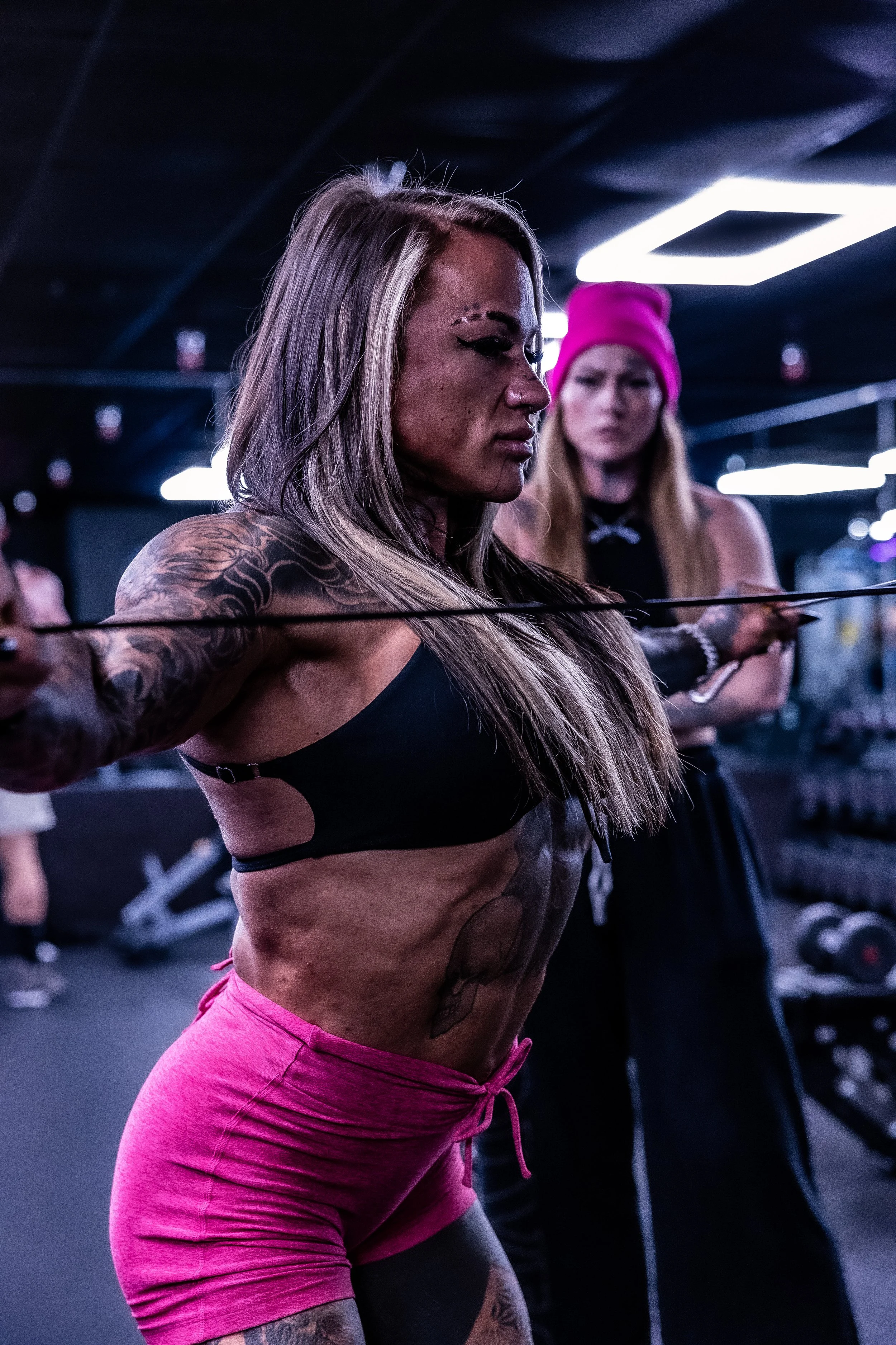 A woman with tattoos on her arm and a serious expression, lifting a barbell during a workout at a gym with black walls and bright rectangular ceiling lights, while another woman in a pink beanie watches in the background.