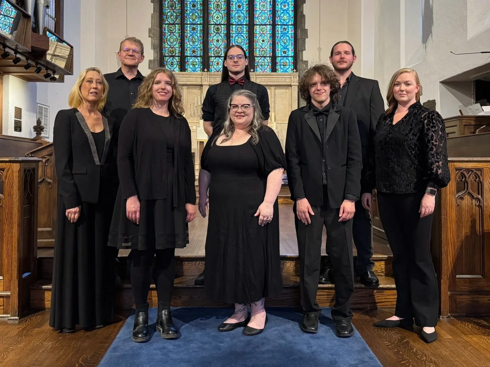 Members of Asheville Vocal Ensemble dressed in formal black attire, standing inside a church with stained glass windows in the background.