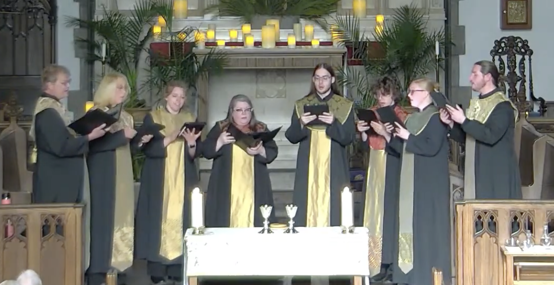 Members of the Asheville Vocal Ensemble standing in a church, holding music, during a performance. The setting includes candles on the altar, potted plants, and ornate woodwork.