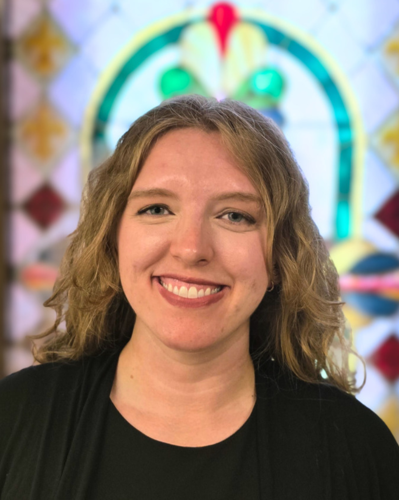 Anna Koogler, Alto. A smiling young woman with wavy blond hair in front of a colorful stained glass window.
