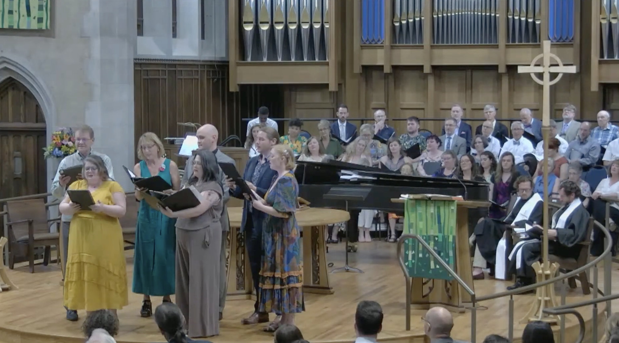 Members of Asheville Vocal Ensemble performing for a wedding with a grand piano on a wooden stage, wooden panels, and a large cross on the back wall. Several choir members are holding music folders.