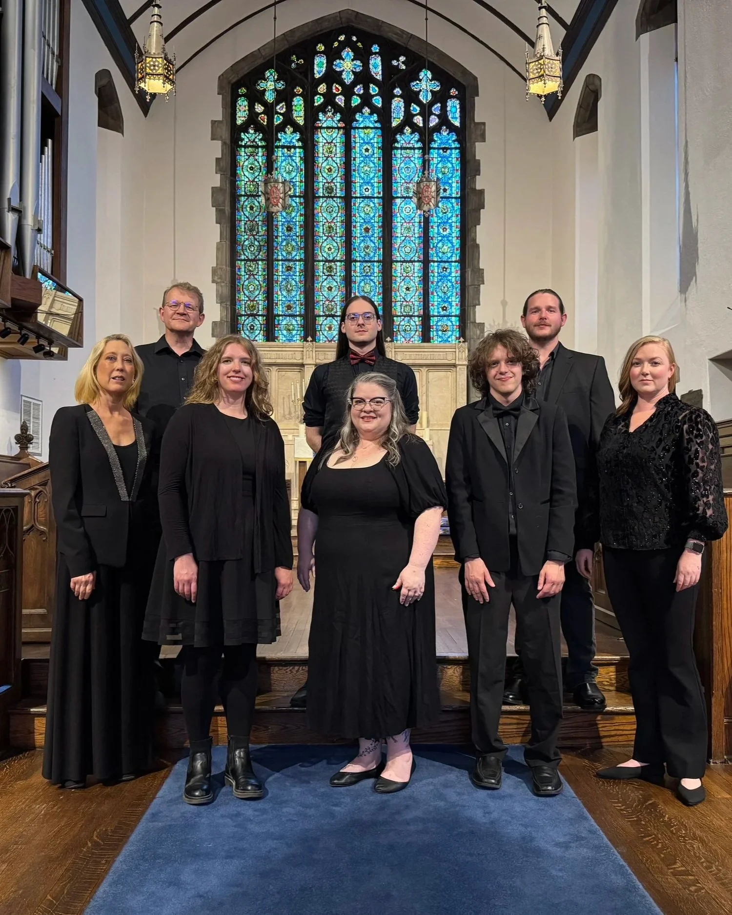 Members of the Asheville Vocal Ensemble in black attire standing on the steps of a church altar with stained glass windows behind them.
