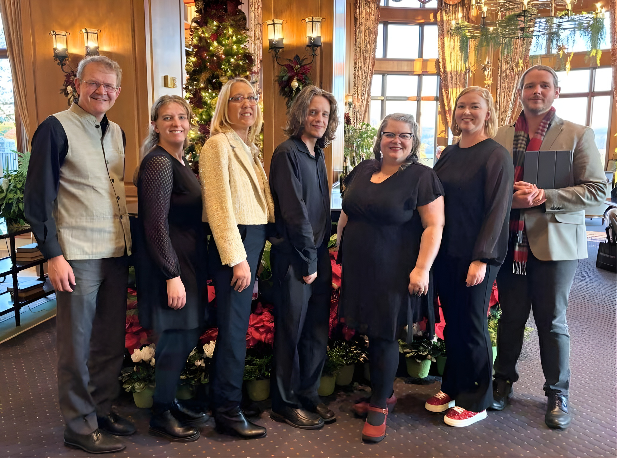 Members of Asheville Vocal Ensemble standing together at the Biltmore Inn in front of a decorated Christmas tree and holiday ornaments, smiling at the camera.