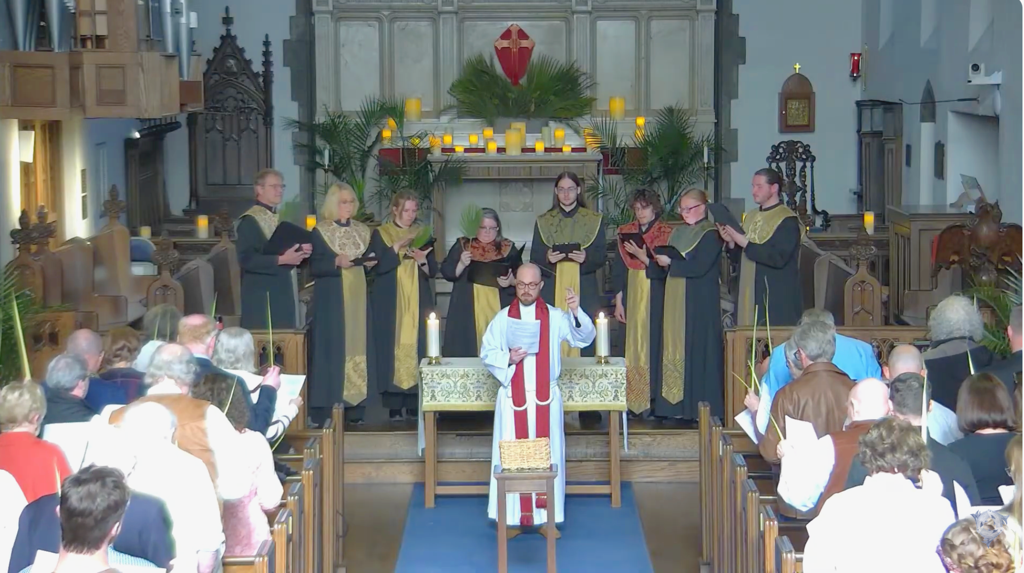 A church congregation during a service with individuals in robes standing at the altar, holding songbooks, and a clergy member in the foreground with a red stole and papers. The church interior has candles, palm fronds, and religious symbols, with attendees seated in wooden pews.