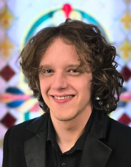 Will Rhodarmer, Baritone. A young man with curly brown hair smiling, wearing a black suit with a black shirt, standing in front of a colorful, blurred background.