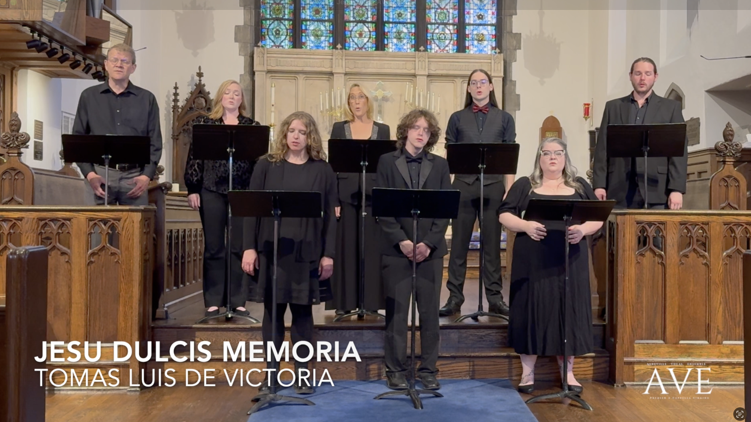 Members of the Asheville Vocal Ensemble performing inside a church with stained glass windows and ornate wooden decor. The choir members are dressed in black, standing in front of the altar with music stands.