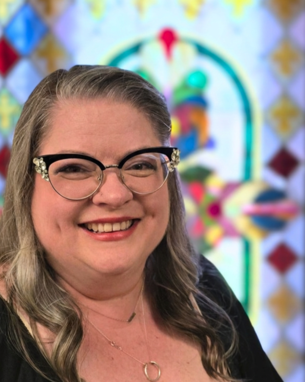 Nana Hosmer Griffin, Soprano / Alto. A woman with shoulder-length, brown hair, wearing round glasses with floral details on the corners. The background features colorful stained glass art.