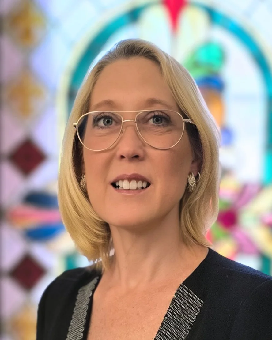 Pamela Miller, Soprano. A woman with blonde hair, glasses, and earrings, smiling in front of a colorful, blurred background.