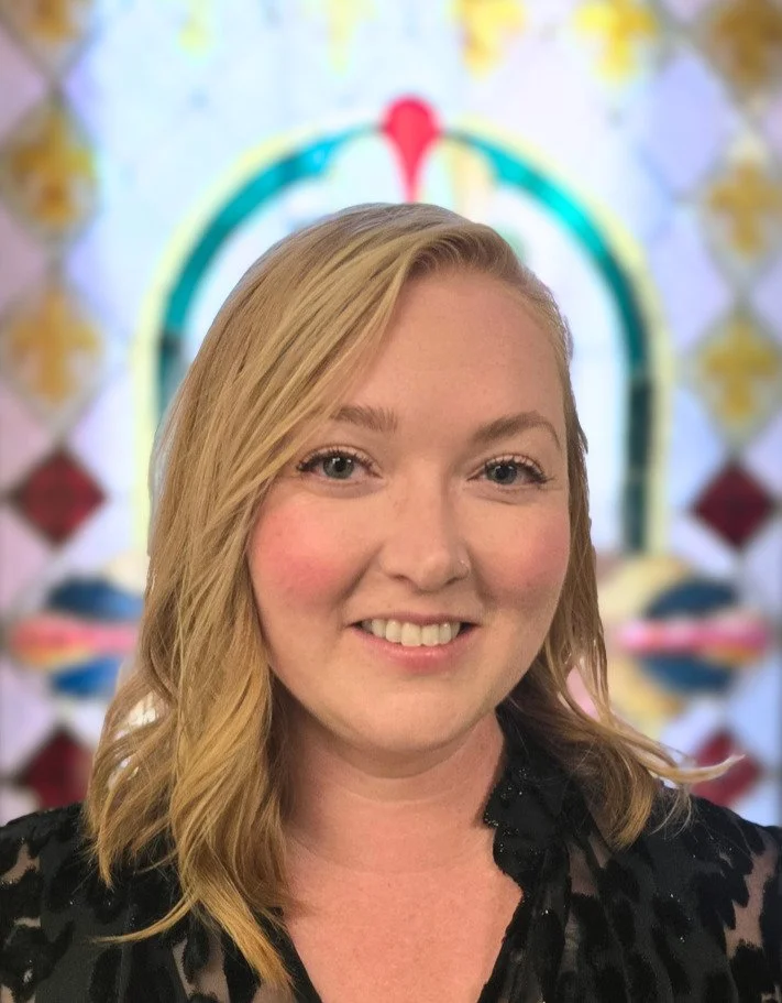 Caitlin Nelson, Soprano / Alto. A woman with strawberry blonde hair and a black top smiling in front of a colorful, blurred background.