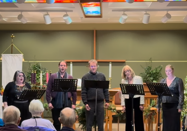 Members of Asheville Vocal Ensemble singing on stage in front of an audience, with music stands and Christmas decorations.