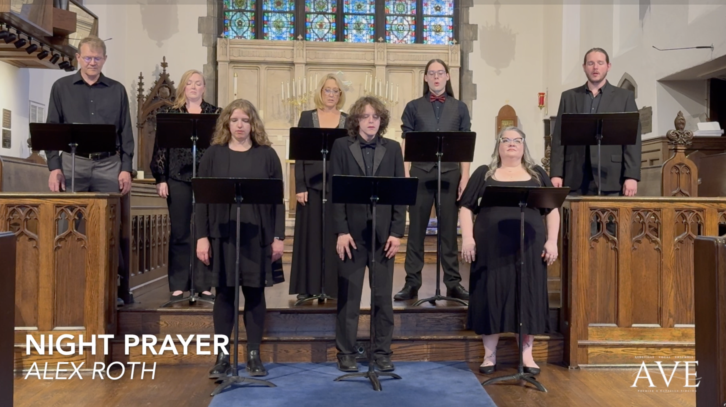 Members of the Asheville Vocal Ensemble standing and singing in a church.  The background shows stained glass windows and a wooden altar.