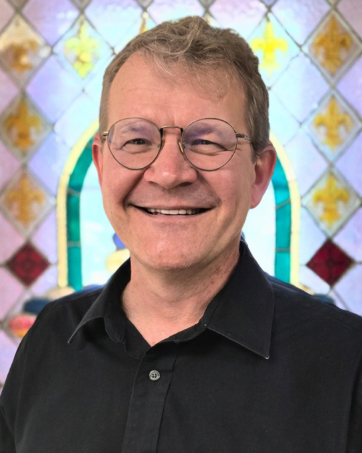 Jeff Konz, Tenor. A smiling man with glasses and short, light brown hair, wearing a black collared shirt, posing in front of a colorful stained glass window.