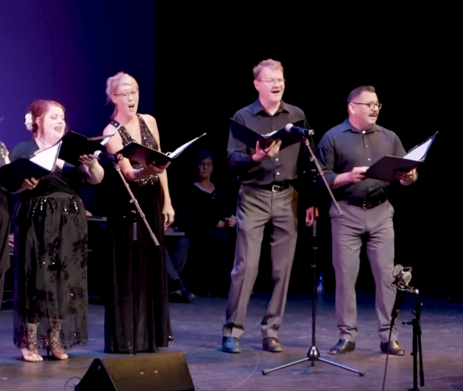 Members of Asheville Vocal Ensemble performing on stage, holding sheet music, with microphones set up in front of them.
