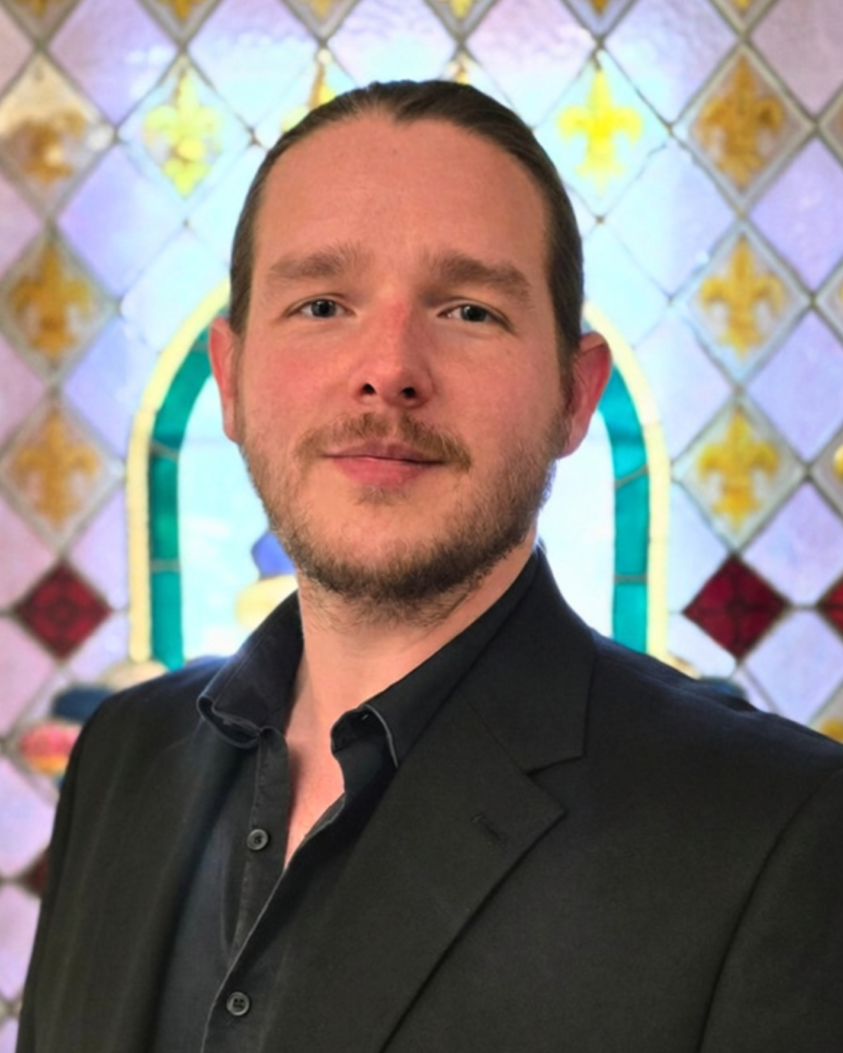 Casey Edwards, Tenor. A man with light skin, brown hair, and a beard wearing a black suit jacket and black shirt, standing in front of a stained-glass background in a church or similar setting.